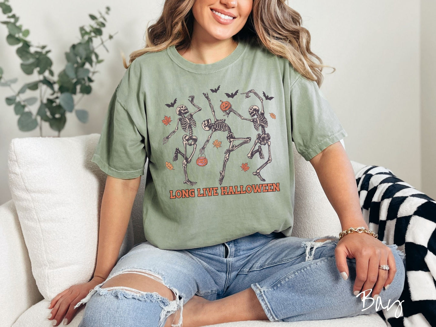 A woman wearing a cute, vintage bay colored Comfort Colors T-shirt with the text Long Live Halloween in orange font. Above are three dancing skeletons holding pumpkins and jackolantern trick or treat buckets with bats and leaves in the air.