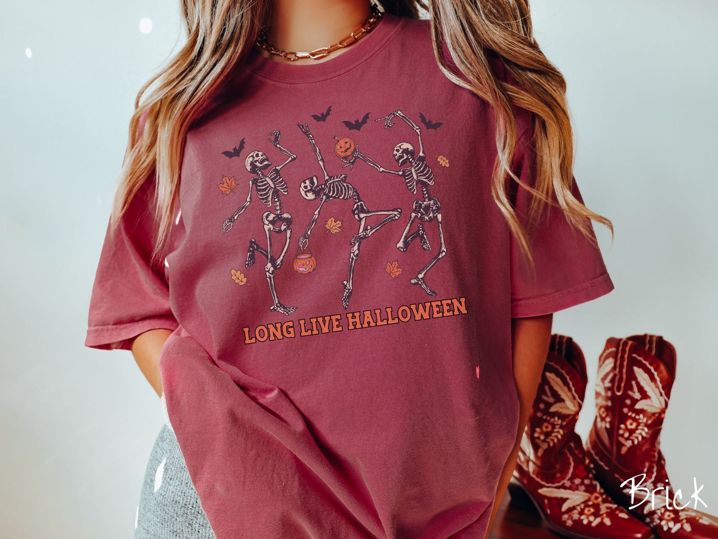A woman wearing a cute, vintage brick colored Comfort Colors T-shirt with the text Long Live Halloween in orange font. Above are three dancing skeletons holding pumpkins and jackolantern trick or treat buckets with bats and leaves in the air.