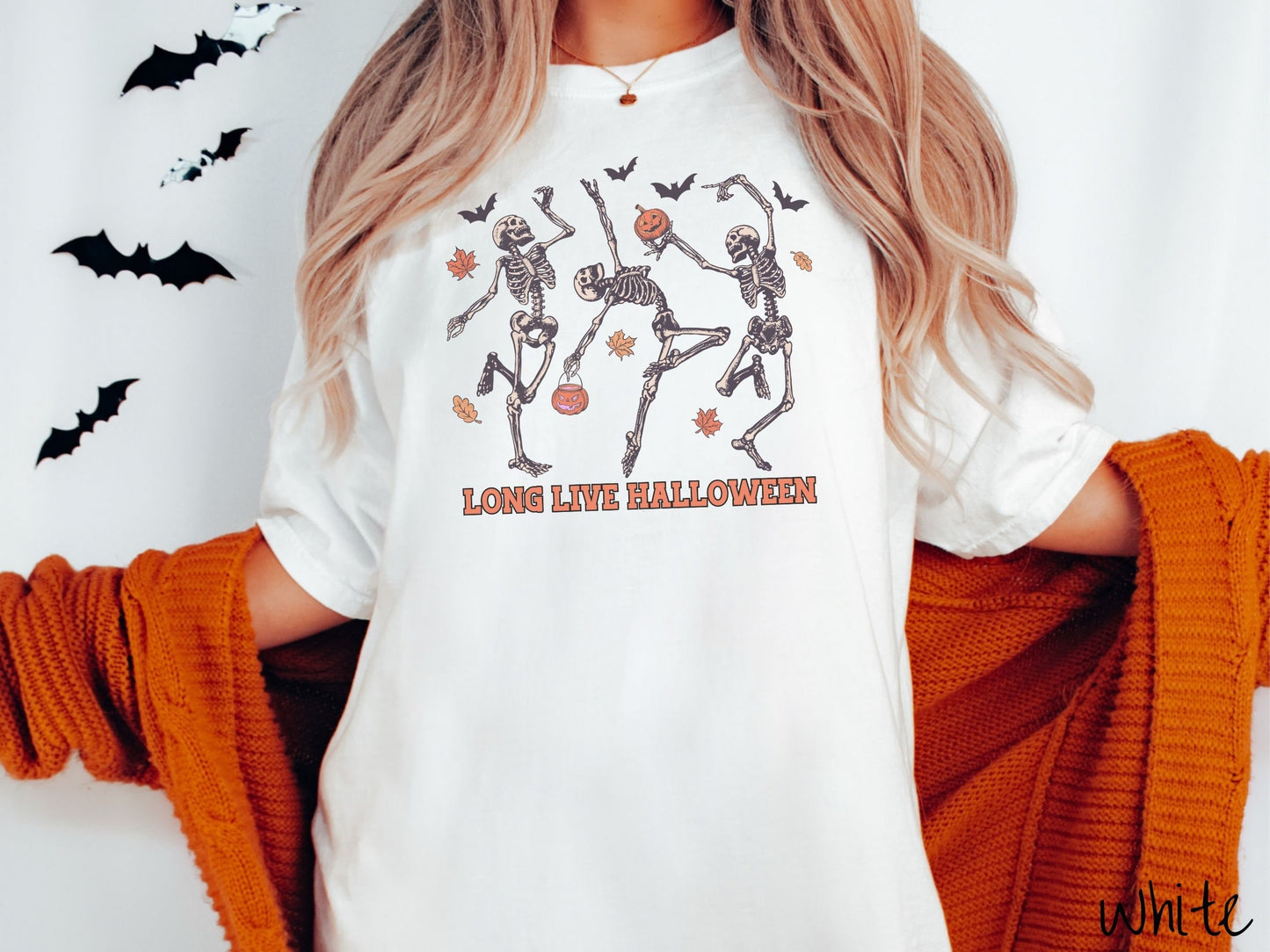 A woman wearing a cute, vintage white colored Comfort Colors T-shirt with the text Long Live Halloween in orange font. Above are three dancing skeletons holding pumpkins and jackolantern trick or treat buckets with bats and leaves in the air.