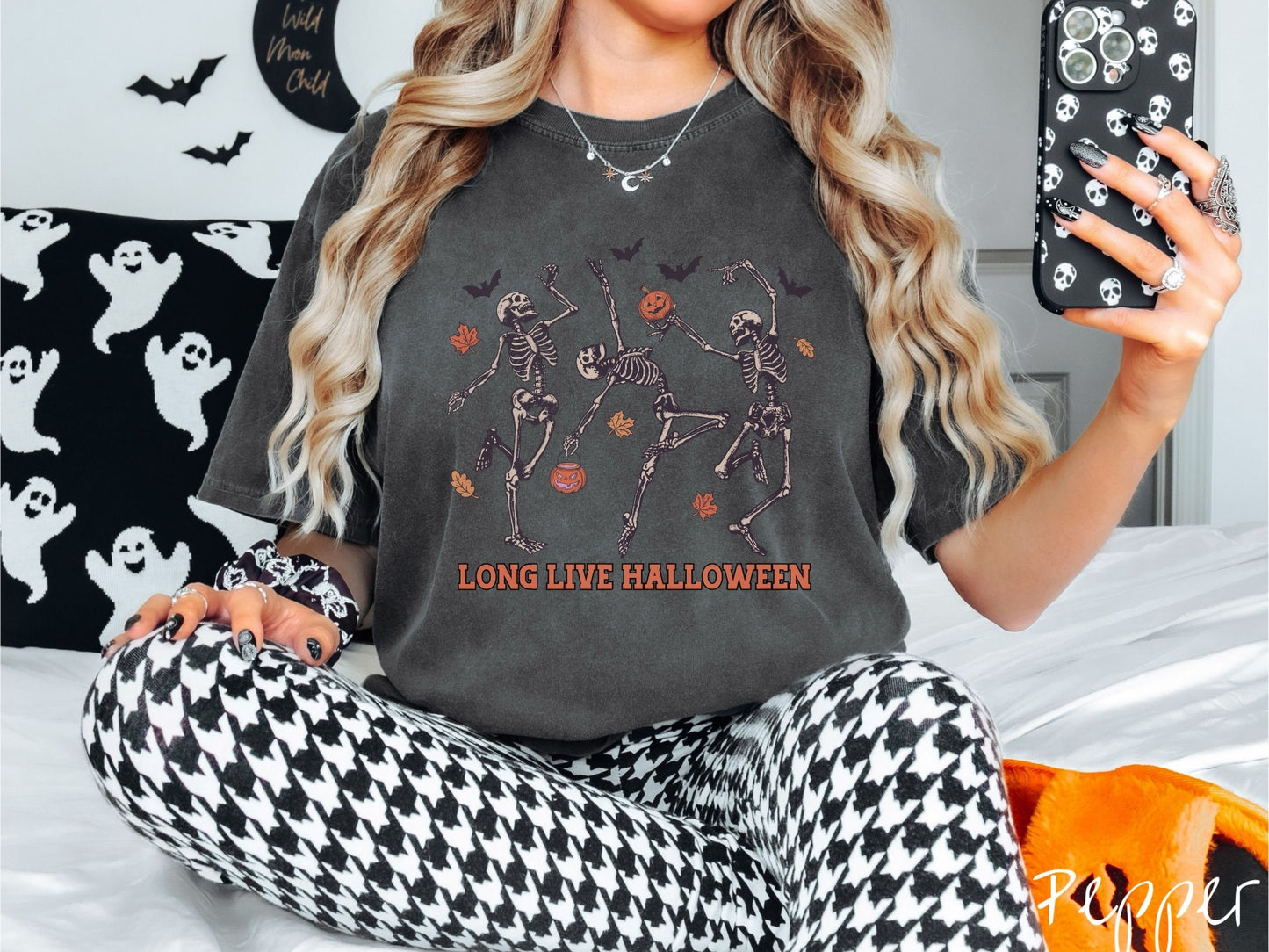 A woman wearing a cute, vintage pepper colored Comfort Colors T-shirt with the text Long Live Halloween in orange font. Above are three dancing skeletons holding pumpkins and jackolantern trick or treat buckets with bats and leaves in the air.