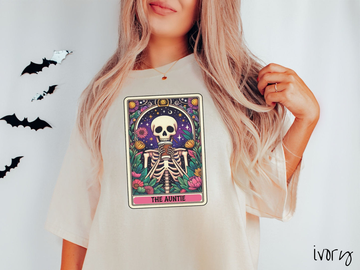 A woman wearing a cute, vintage ivory colored Comfort Colors T-shirt with text The Auntie along the bottom of a tarot card. Above is a smiling skeleton holding a to-go cup of coffee up to her mouth, and is surrounded by stars and colorful flowers.