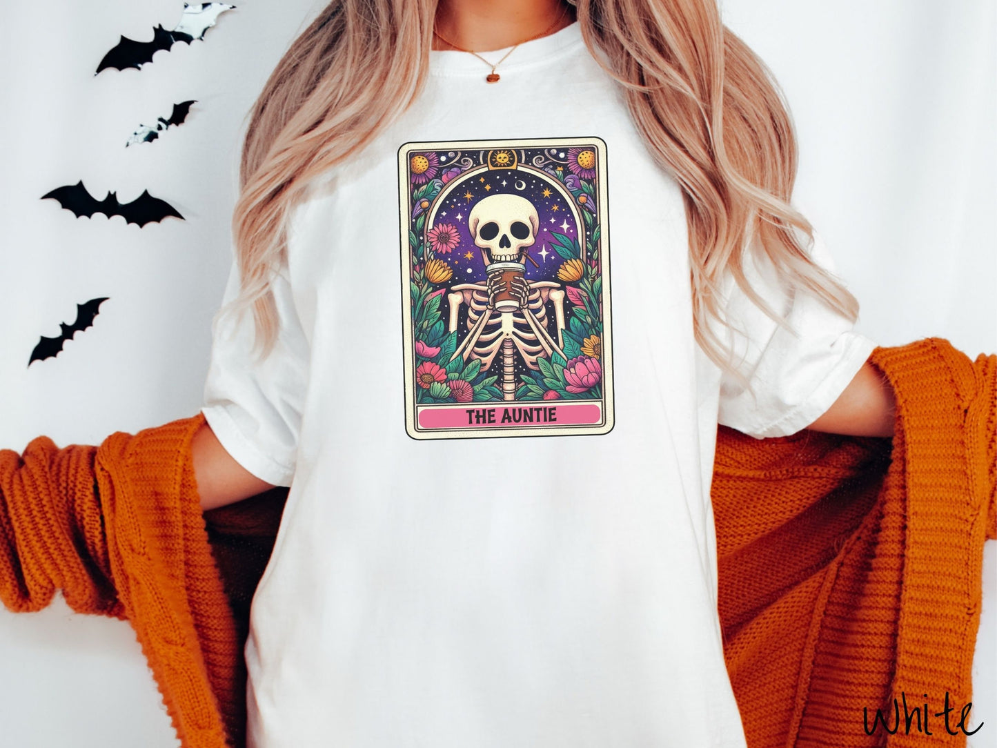 A woman wearing a cute, vintage white colored Comfort Colors T-shirt with text The Auntie along the bottom of a tarot card. Above is a smiling skeleton holding a to-go cup of coffee up to her mouth, and is surrounded by stars and colorful flowers.