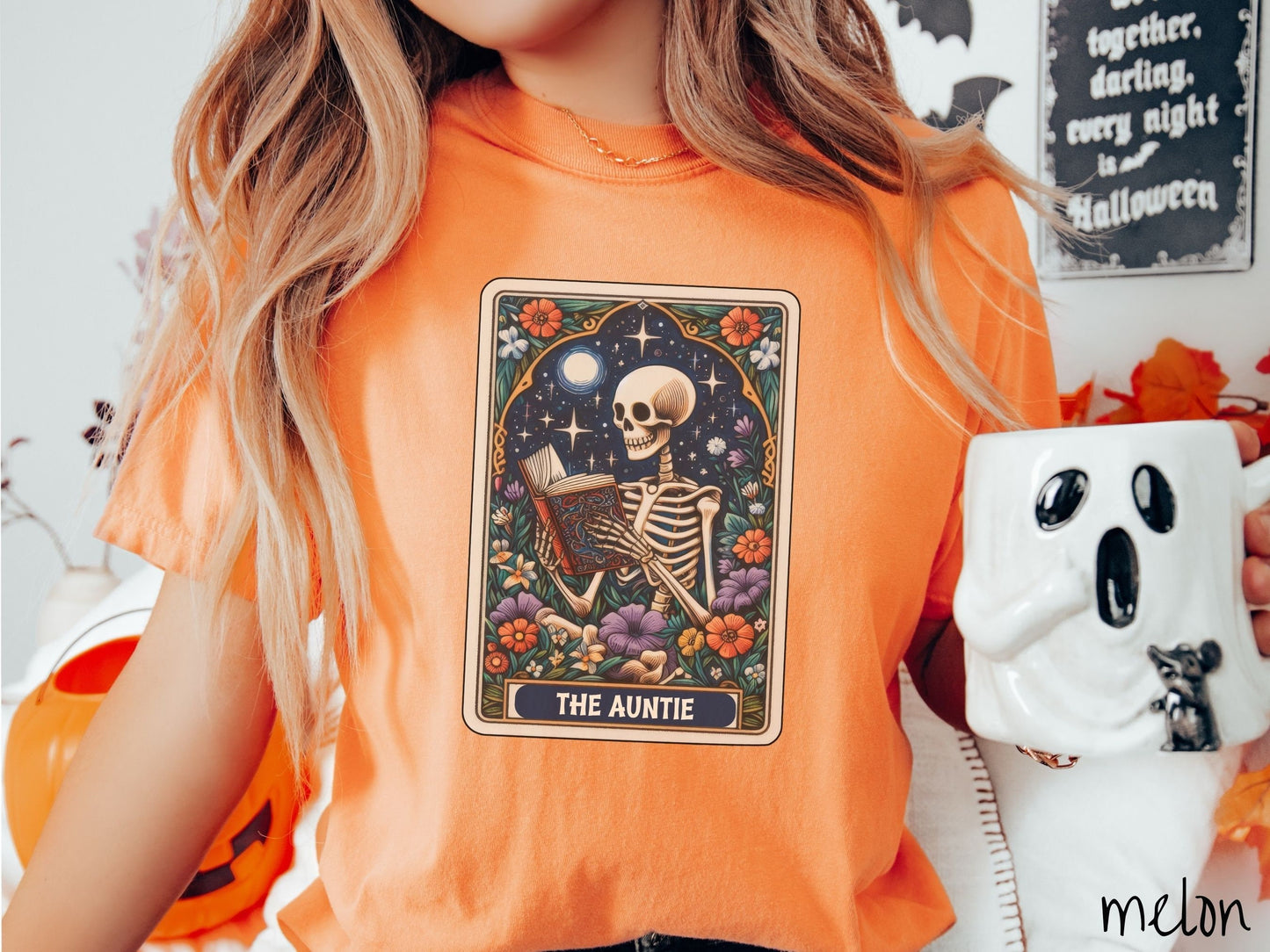A woman wearing a cute, vintage melon colored Comfort Colors T-shirt with text The Auntie along the bottom of a tarot card. Above is a skeleton reading an old looking book while under the moon and stars and surrounded by colorful flowers.