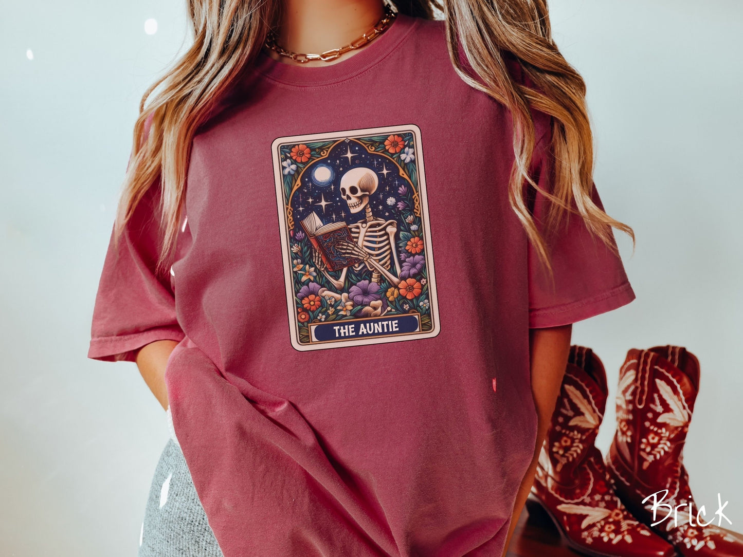 A woman wearing a cute, vintage brick colored Comfort Colors T-shirt with text The Auntie along the bottom of a tarot card. Above is a skeleton reading an old looking book while under the moon and stars and surrounded by colorful flowers.
