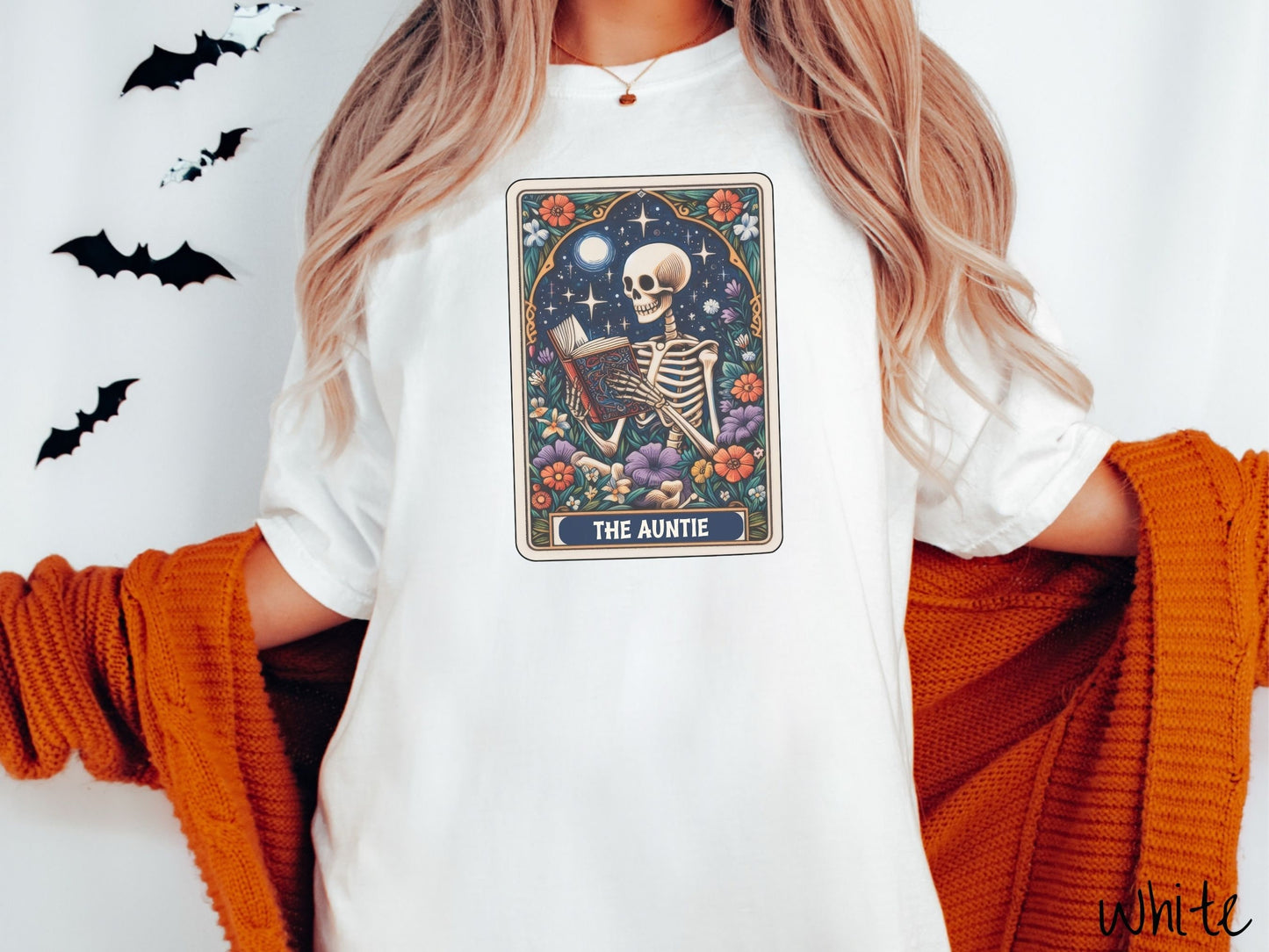 A woman wearing a cute, vintage white colored Comfort Colors T-shirt with text The Auntie along the bottom of a tarot card. Above is a skeleton reading an old looking book while under the moon and stars and surrounded by colorful flowers.