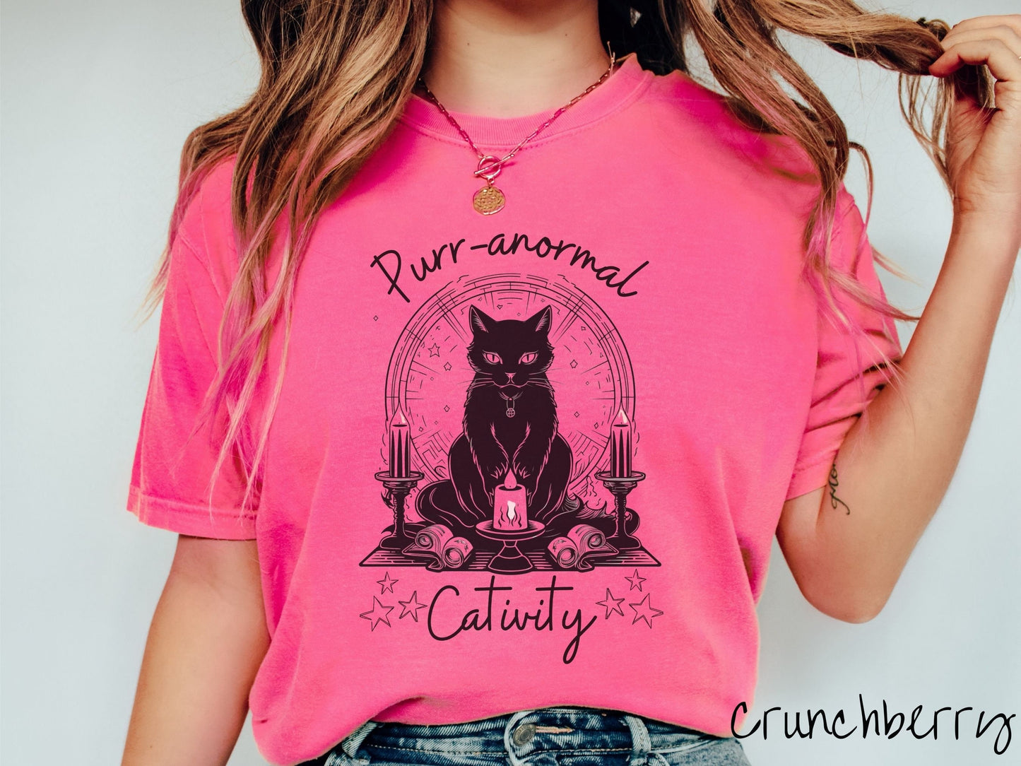 A woman wearing a cute, vintage crunchberry colored Comfort Colors T-shirt with text Purr-anormal Cativity in black font. There is a black cat sitting on an altar with candles and scrolls, with piercing eyes and wearing a necklace.