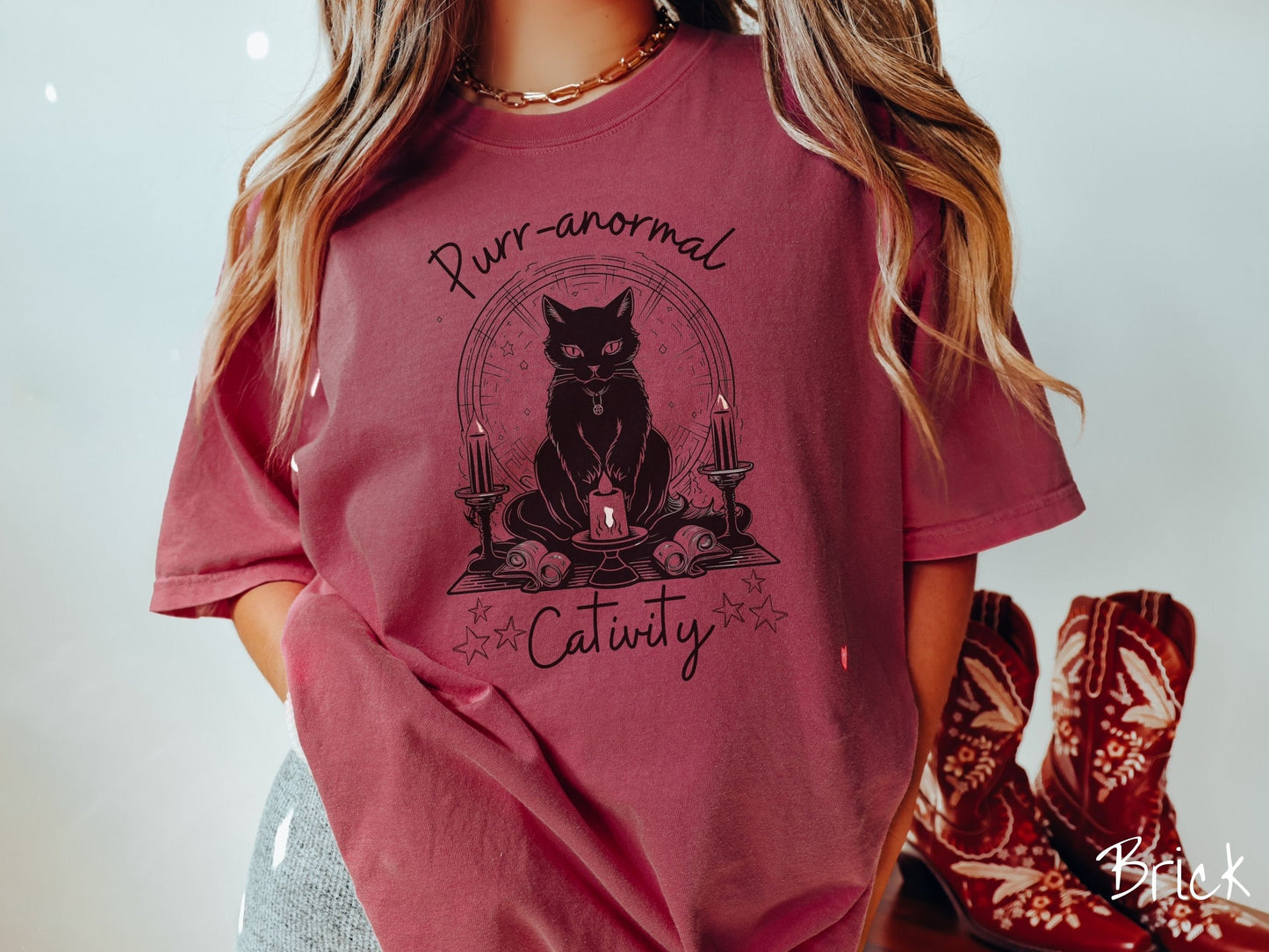 A woman wearing a cute, vintage brick colored Comfort Colors T-shirt with text Purr-anormal Cativity in black font. There is a black cat sitting on an altar with candles and scrolls, staring straight forward with piercing eyes and wearing a necklace.