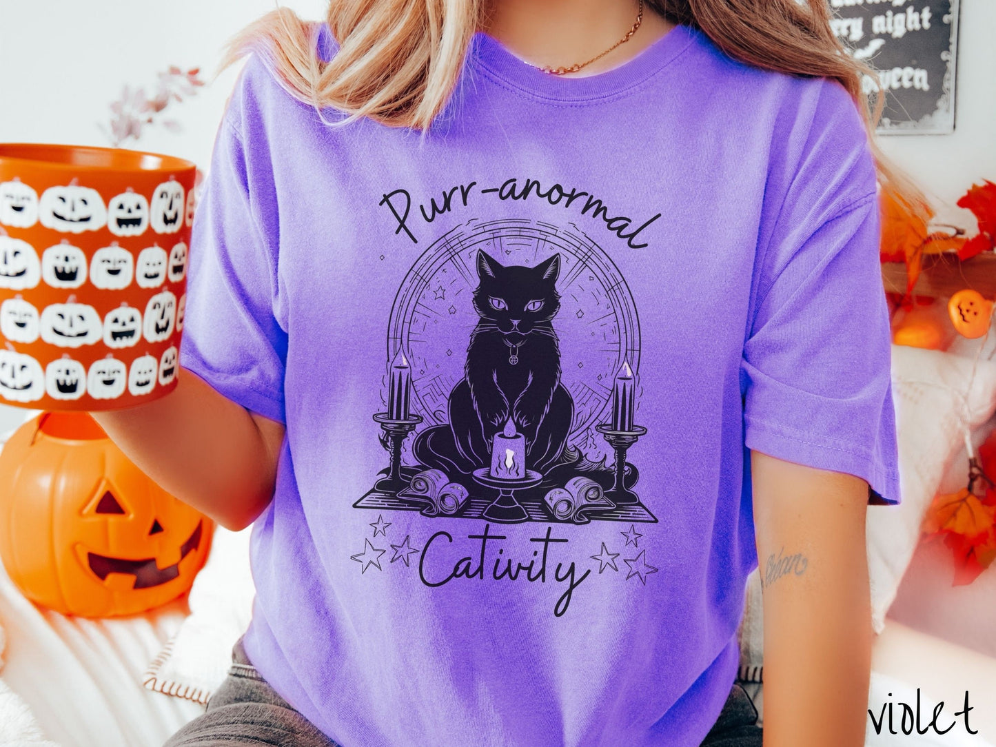 A woman wearing a cute, vintage violet colored Comfort Colors T-shirt with text Purr-anormal Cativity in black font. There is a black cat sitting on an altar with candles and scrolls, staring forward with piercing eyes and wearing a necklace.