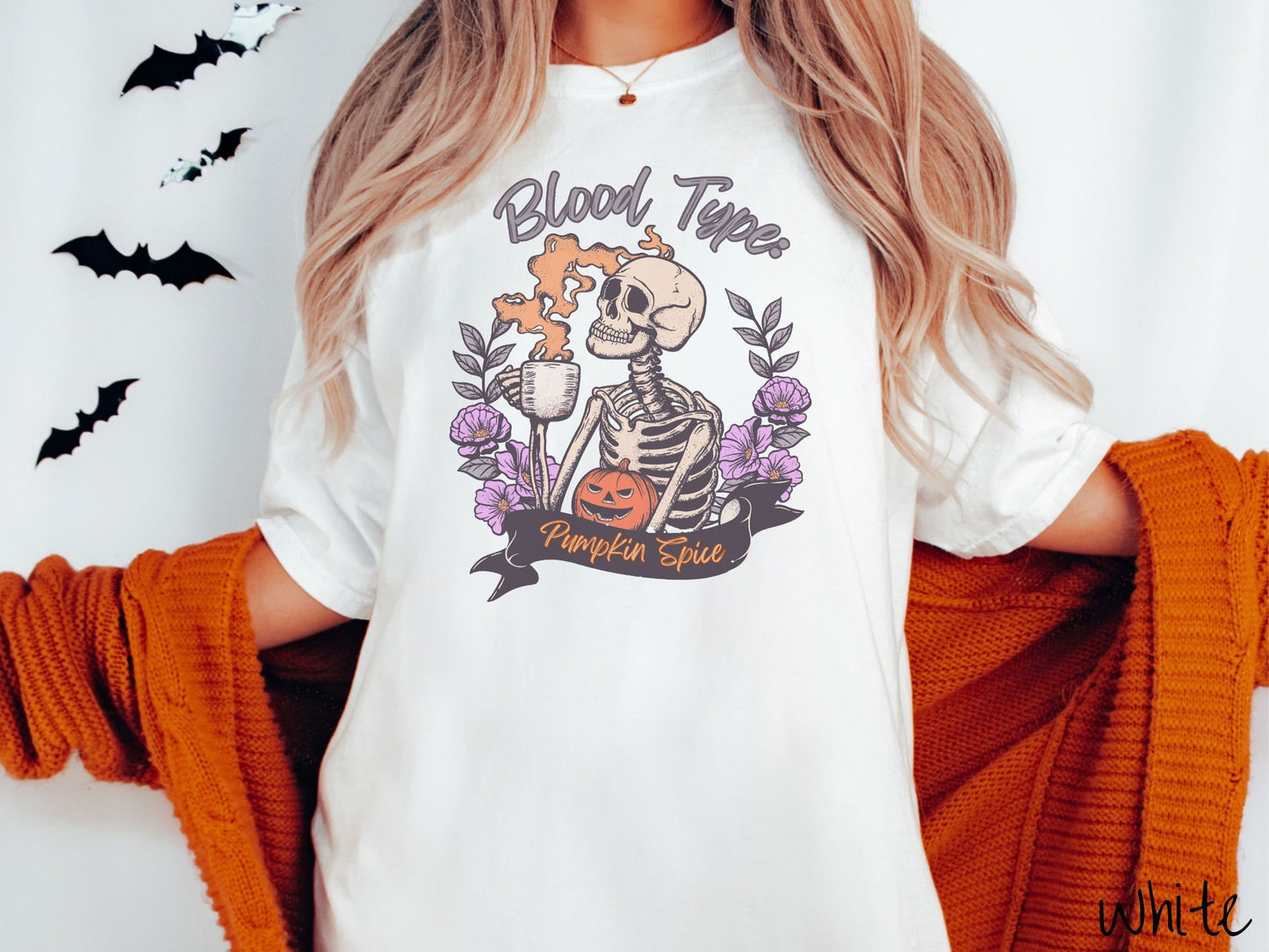 A woman wearing a cute, vintage white colored Comfort Colors T-shirt with text Blood Type: Pumpkin Spice. There is a skeleton holding a pumpkin spiced latte with orange steam coming out and a pumpkin while surrounded by purple flowers.
