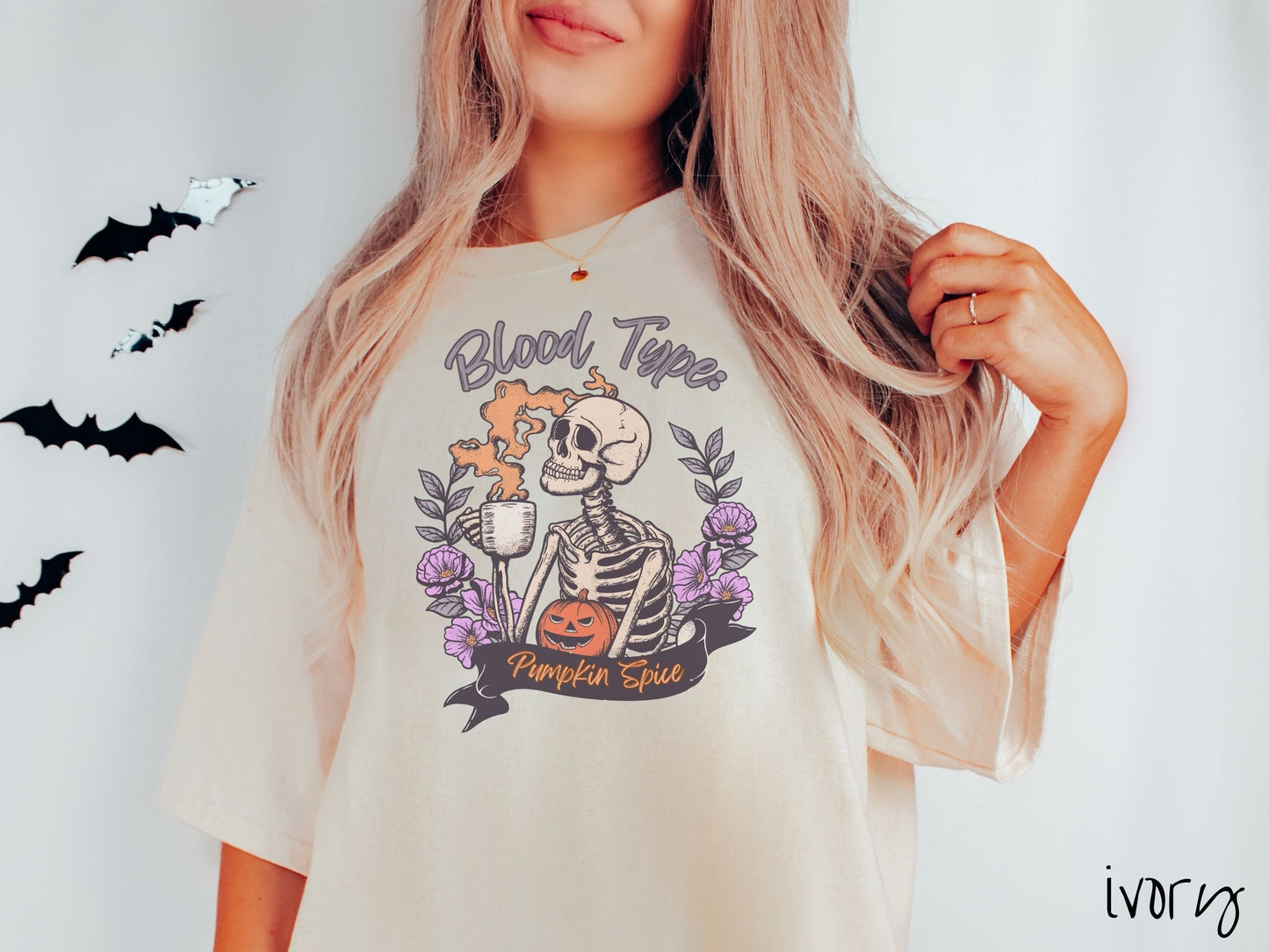 A woman wearing a cute, vintage ivory colored Comfort Colors T-shirt with text Blood Type: Pumpkin Spice. There is a skeleton holding a pumpkin spiced latte with orange steam coming out and a pumpkin while surrounded by purple flowers.