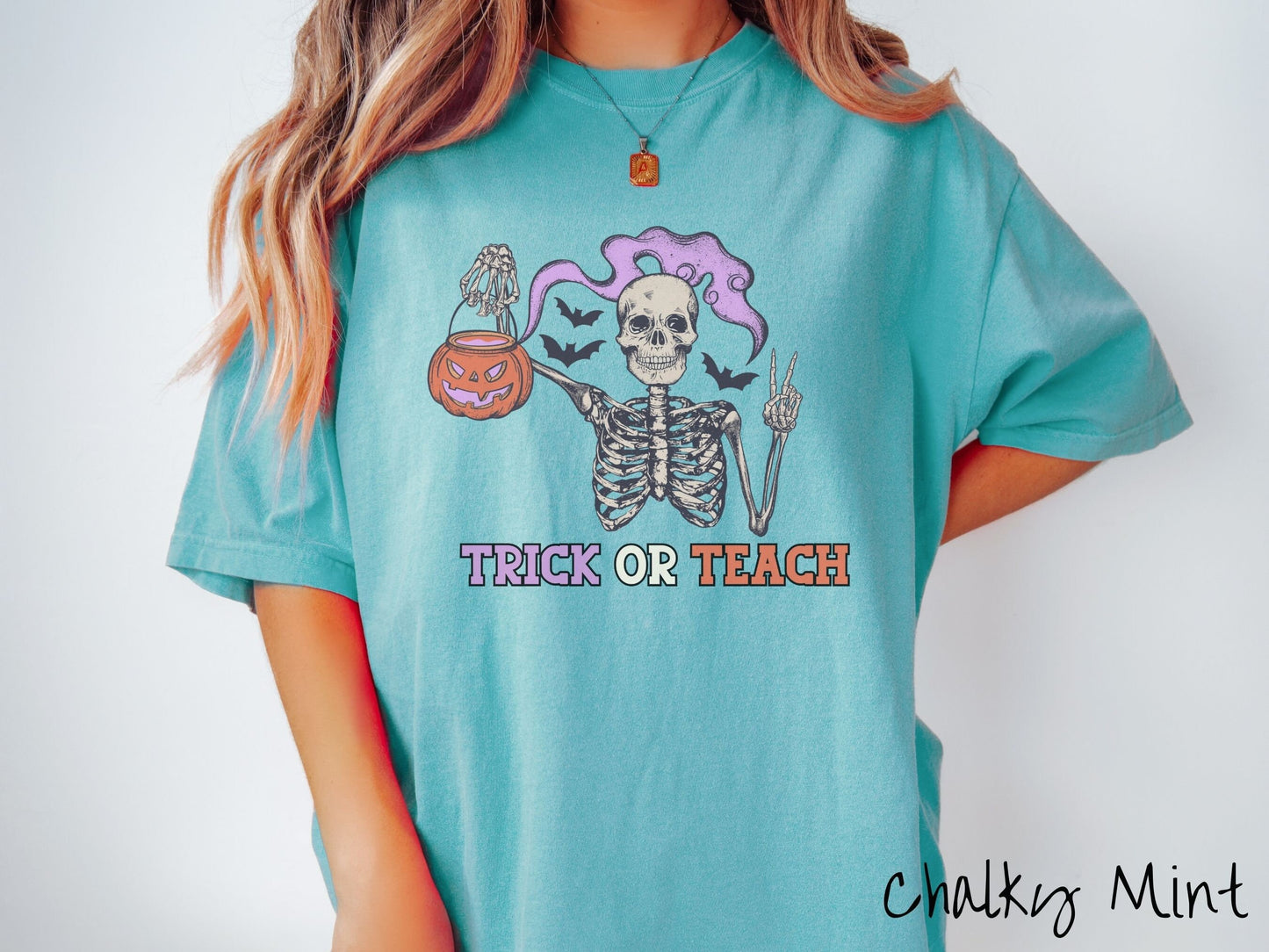 A woman wearing a cute, vintage chalky mint colored Comfort Colors T-shirt with text Trick or Teach in purple and orange font. Above is a skeleton holding up a peace sign and a pumpkin candy bucket with purple smoke coming out and bats in the back.