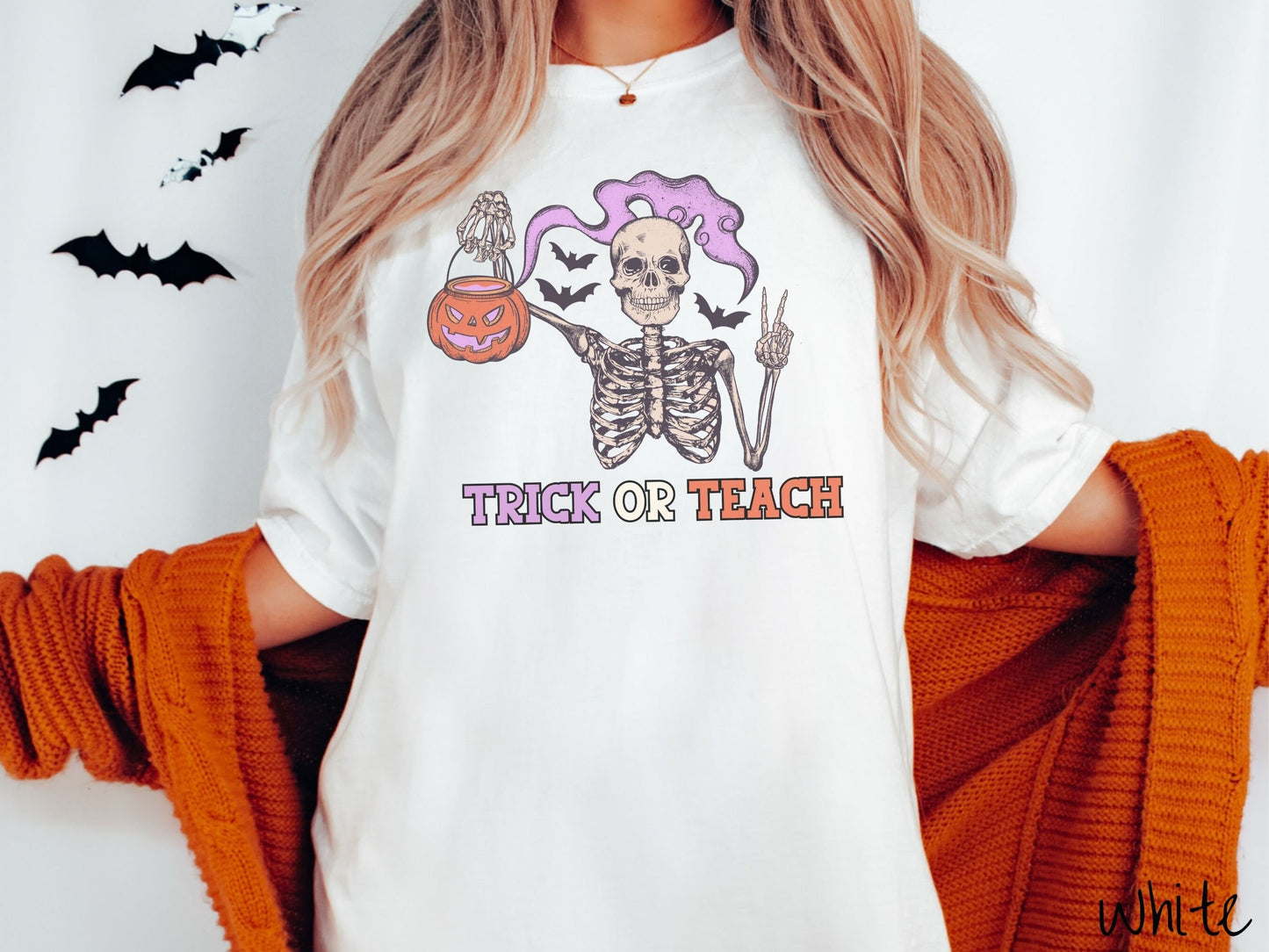 A woman wearing a cute, vintage white colored Comfort Colors T-shirt with text Trick or Teach in purple and orange font. Above is a skeleton holding up a peace sign and a pumpkin candy bucket with purple smoke coming out and bats in the background.