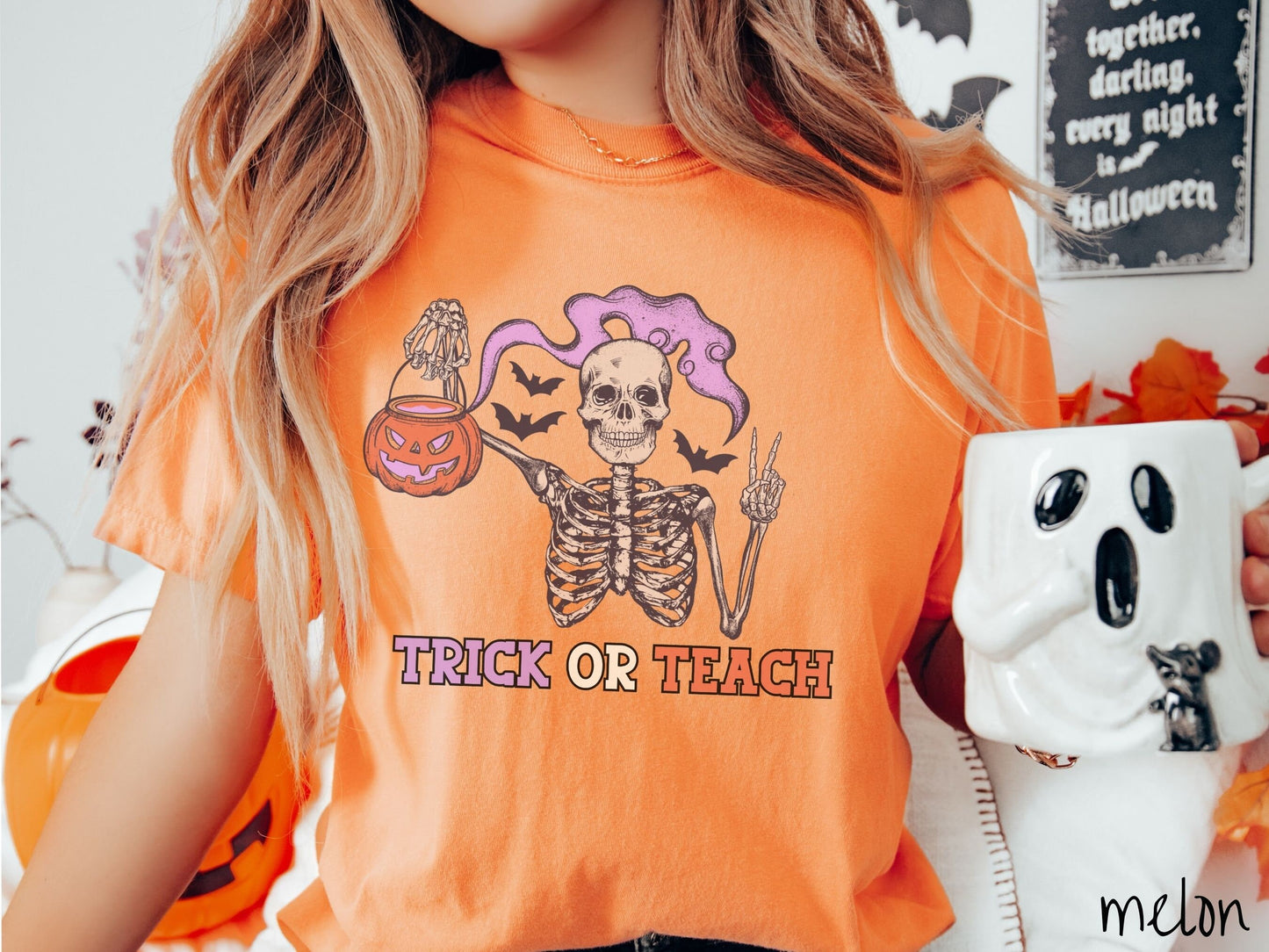 A woman wearing a cute, vintage melon colored Comfort Colors T-shirt with text Trick or Teach in purple and orange font. Above is a skeleton holding up a peace sign and a pumpkin candy bucket with purple smoke coming out and bats in the background.