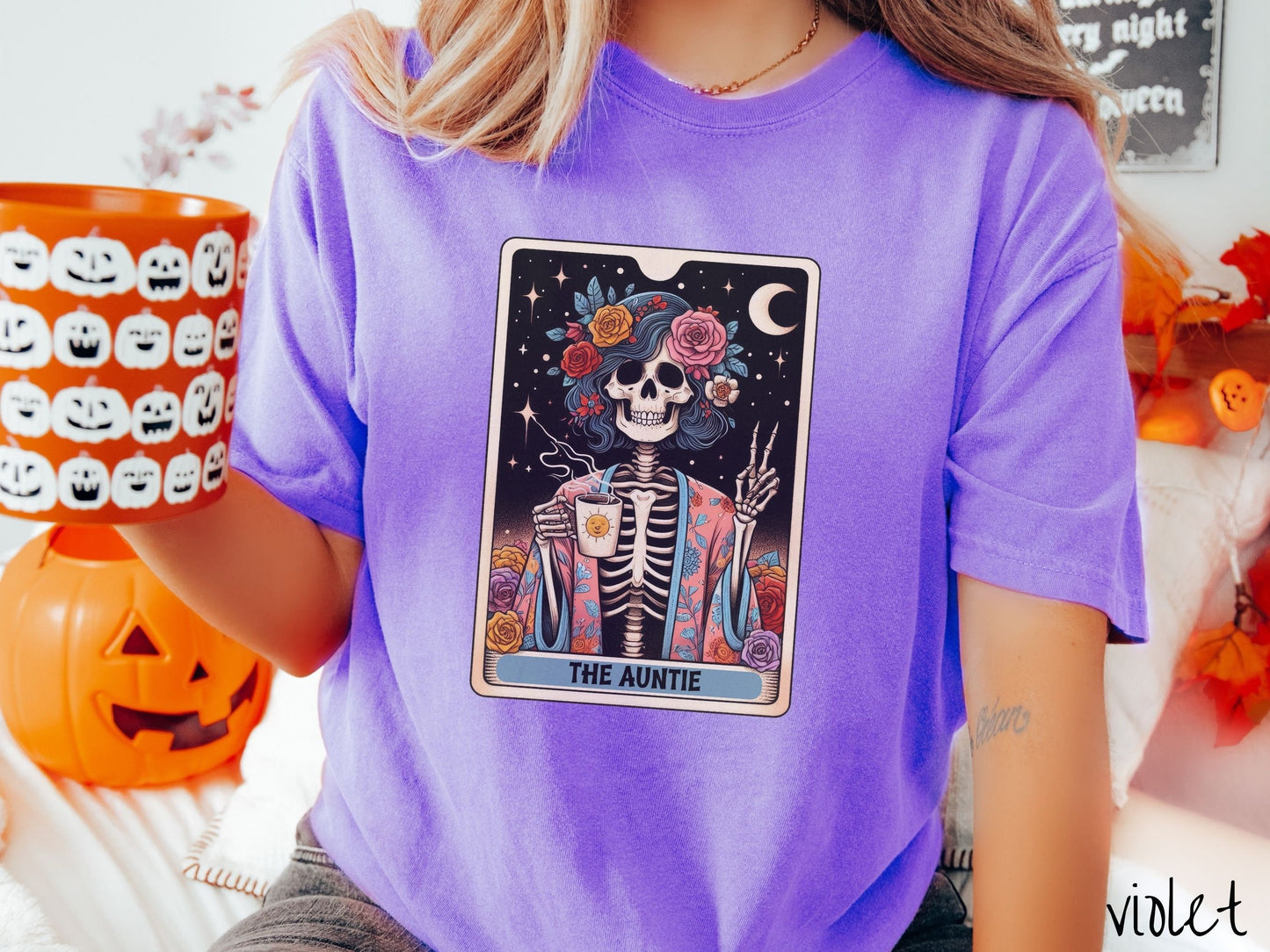 A woman wearing a cute, vintage violet colored Comfort Colors T-shirt with text The Auntie along the bottom of a tarot card. Above is a picture of a female skeleton holding up a peace sign and wearing a flower robe holding a steaming cup of coffee.
