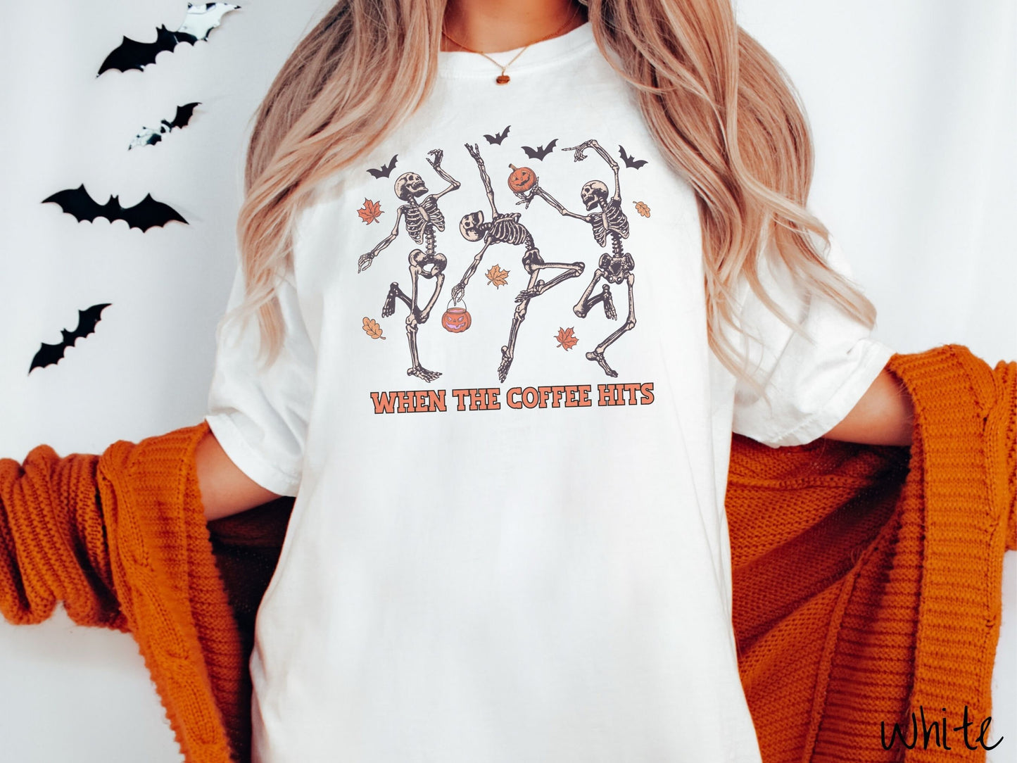 A woman wearing a cute, vintage white colored Comfort Colors T-shirt with the text When the Coffee Hits in orange font. Above are three dancing skeletons holding pumpkins and jackolantern trick or treat buckets with bats and leaves in the air.