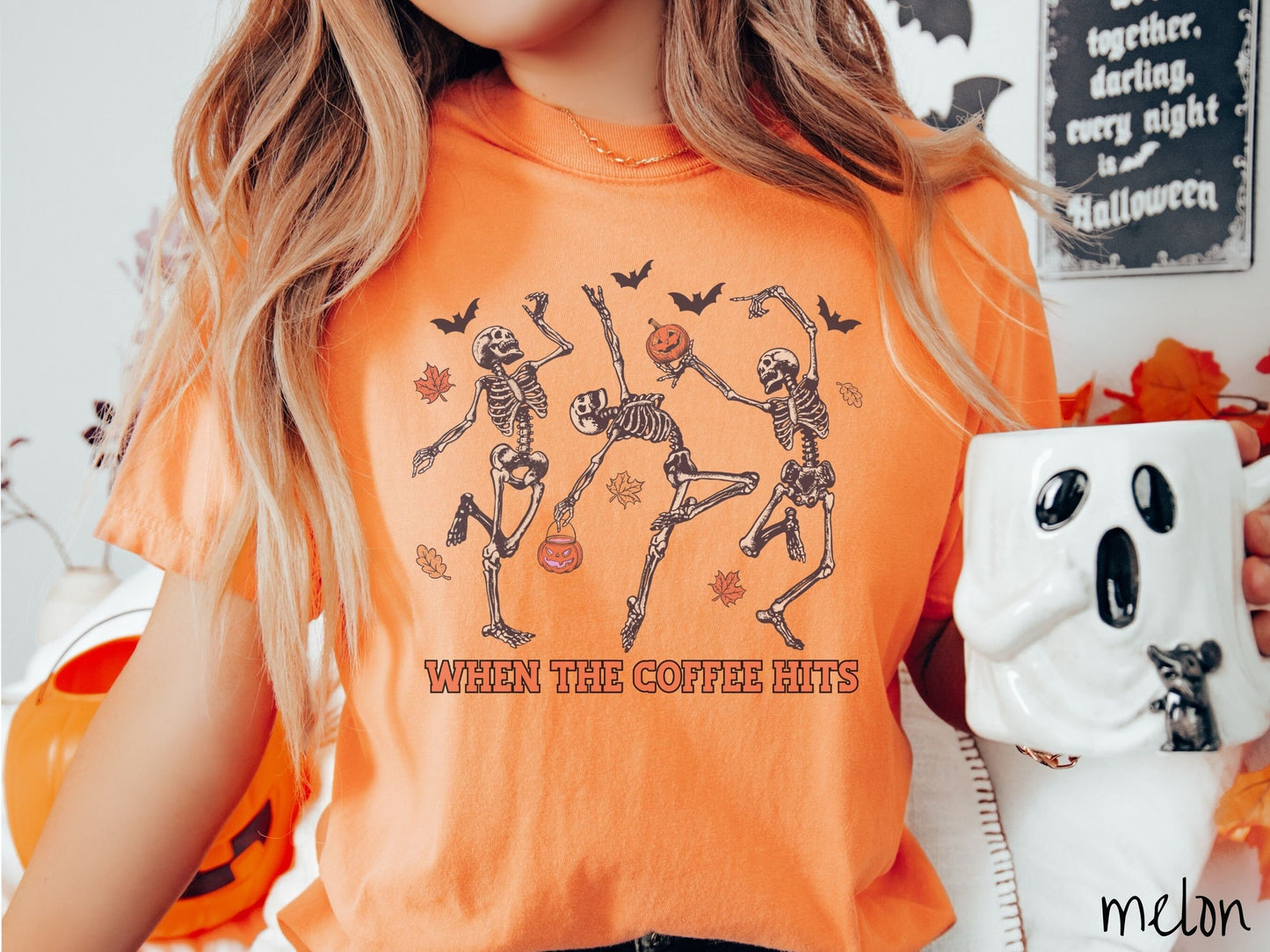 A woman wearing a cute, vintage melon colored Comfort Colors T-shirt with the text When the Coffee Hits in orange font. Above are three dancing skeletons holding pumpkins and jackolantern trick or treat buckets with bats and leaves in the air.