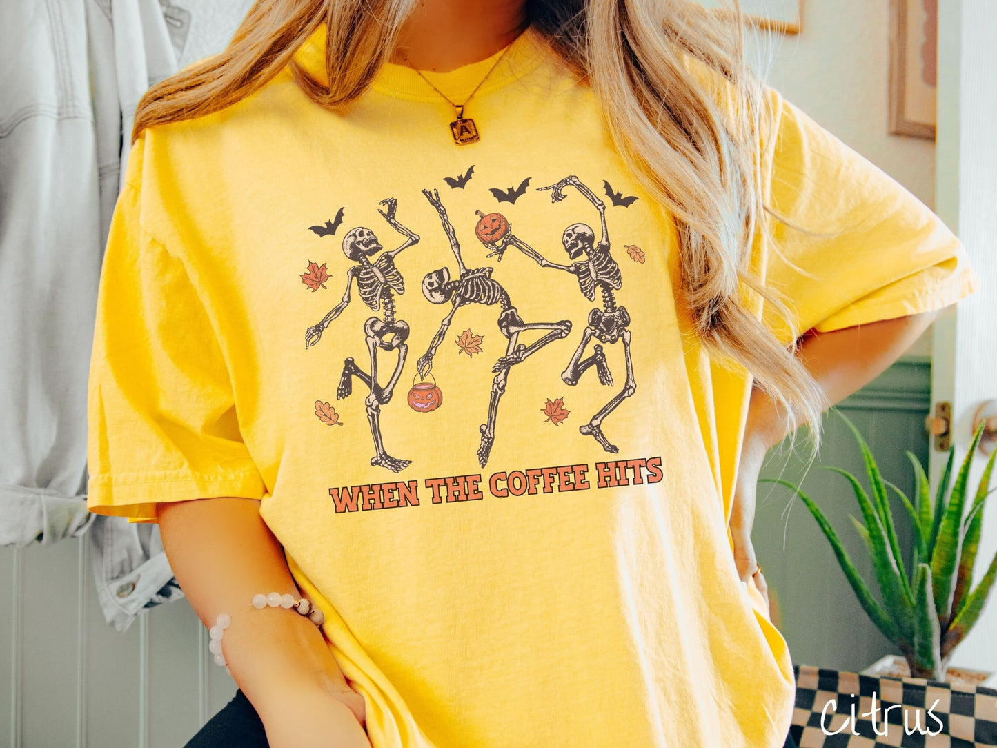 A woman wearing a cute, vintage citrus colored Comfort Colors T-shirt with the text When the Coffee Hits in orange font. Above are three dancing skeletons holding pumpkins and jackolantern trick or treat buckets with bats and leaves in the air.