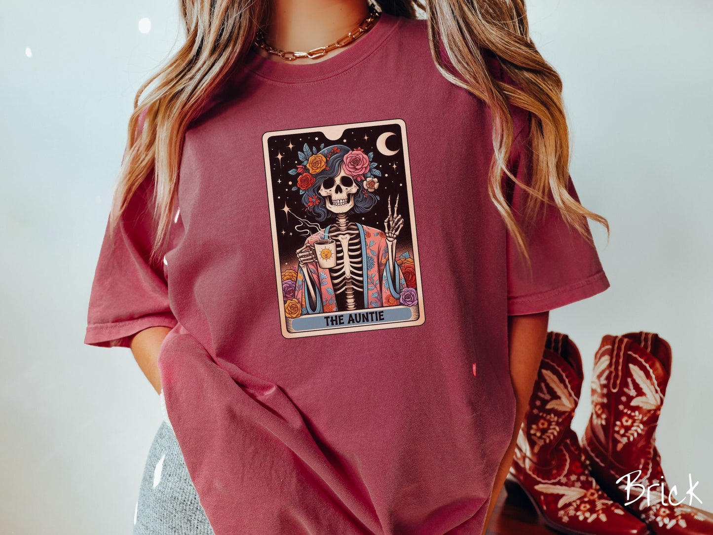 A woman wearing a cute, vintage brick colored Comfort Colors T-shirt with text The Auntie along the bottom of a tarot card. Above is a picture of a female skeleton holding up a peace sign and wearing a flower robe holding a steaming cup of coffee.