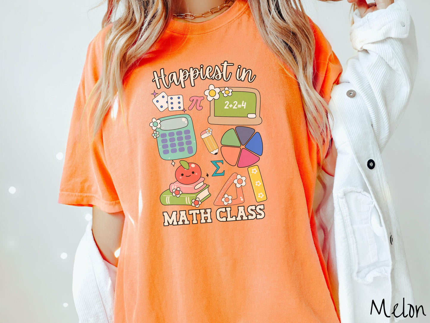 A woman wearing a cute, vintage melon colored Comfort Colors T-shirt with the text Happiest in Math Class. In between the text are the pi symbol, a chalkboard, calculator, pencil, books, protractor, ruler, dice, an apple, and flowers.