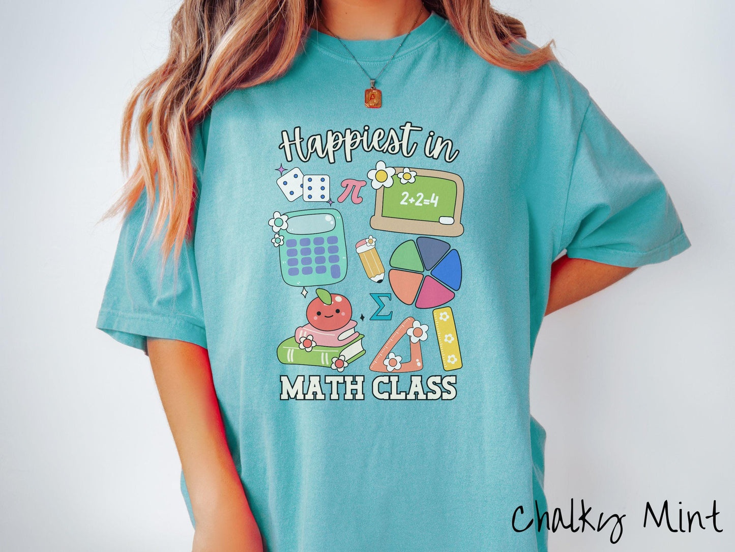 A woman wearing a cute, vintage chalky mint colored Comfort Colors T-shirt with the text Happiest in Math Class. In between the text are the pi symbol, a chalkboard, calculator, pencil, books, protractor, ruler, dice, an apple, and flowers.