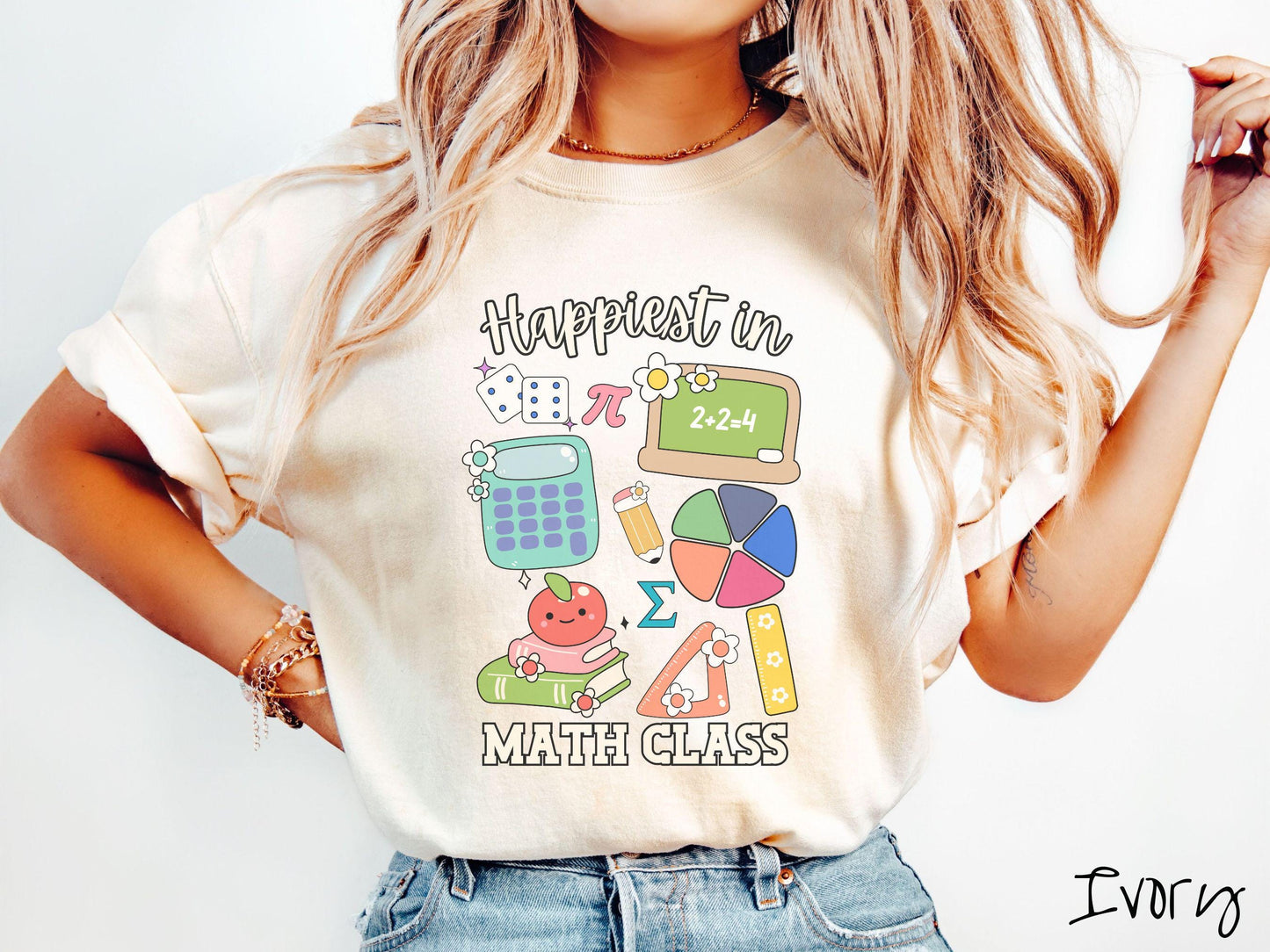 A woman wearing a cute, vintage ivory colored Comfort Colors T-shirt with the text Happiest in Math Class. In between the text are the pi symbol, a chalkboard, calculator, pencil, books, protractor, ruler, dice, an apple, and flowers.