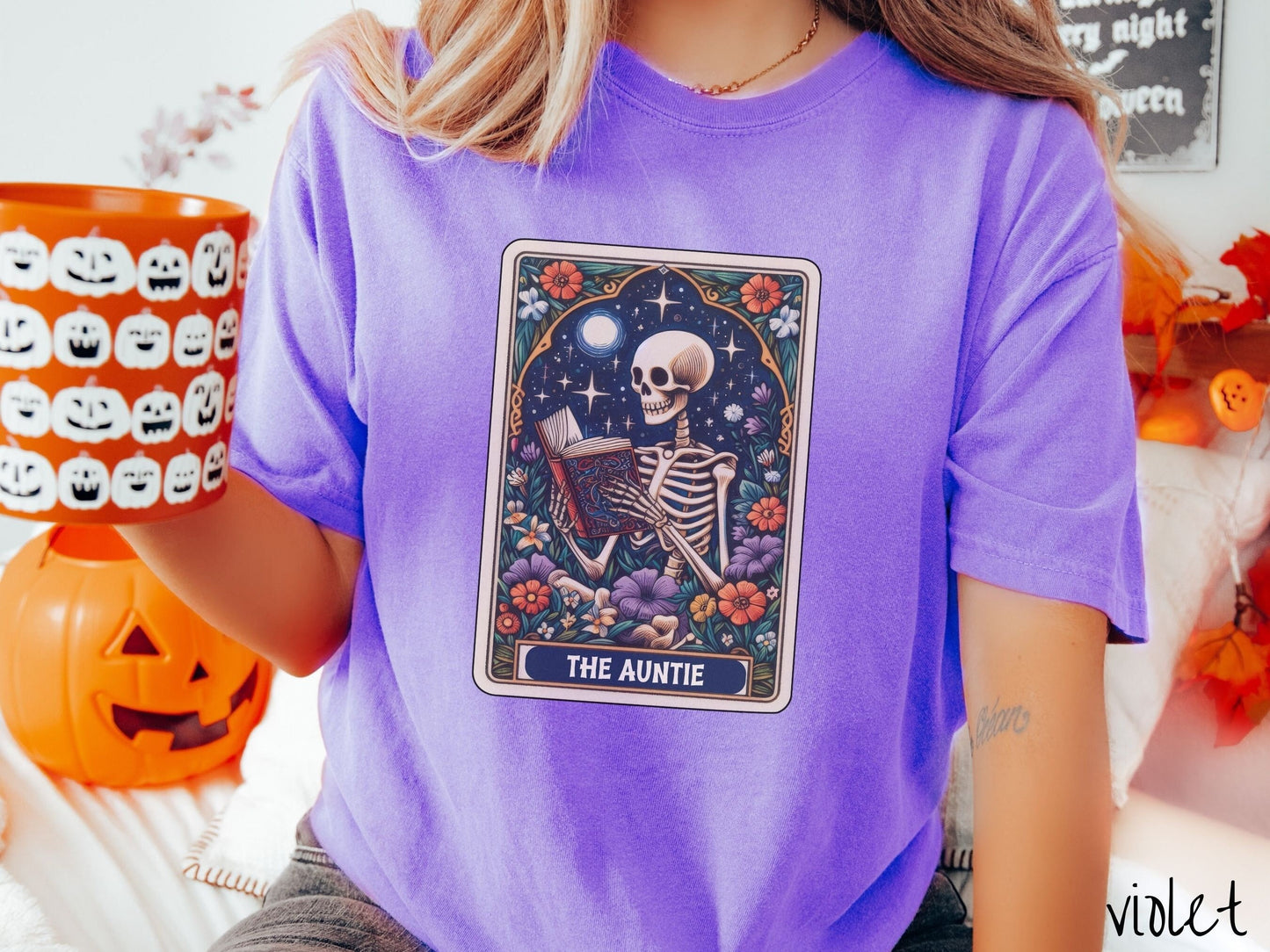 A woman wearing a cute, vintage violet colored Comfort Colors T-shirt with text The Auntie along the bottom of a tarot card. Above is a skeleton reading an old looking book while under the moon and stars and surrounded by colorful flowers.