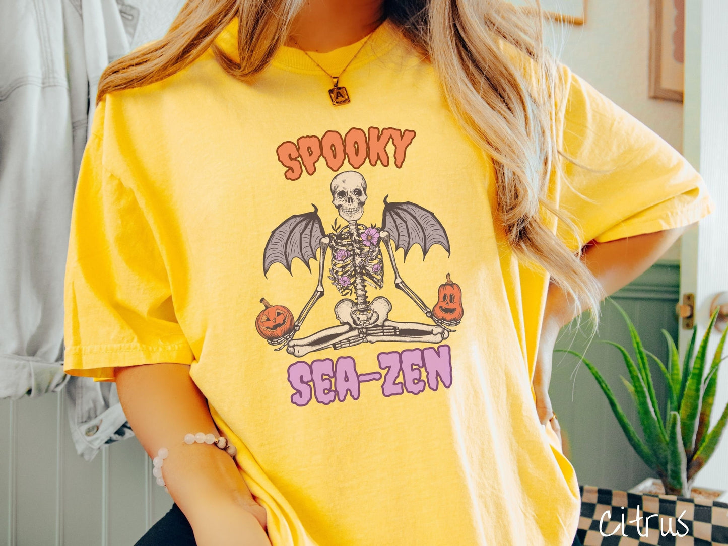 A woman wearing a cute, vintage citrus colored Comfort Colors T-shirt with text Spooky Sea-Zen in orange and purple font. There is a skeleton wearing flowers and bat wings sitting in a full lotus yoga pose while holding pumpkins in both hands.