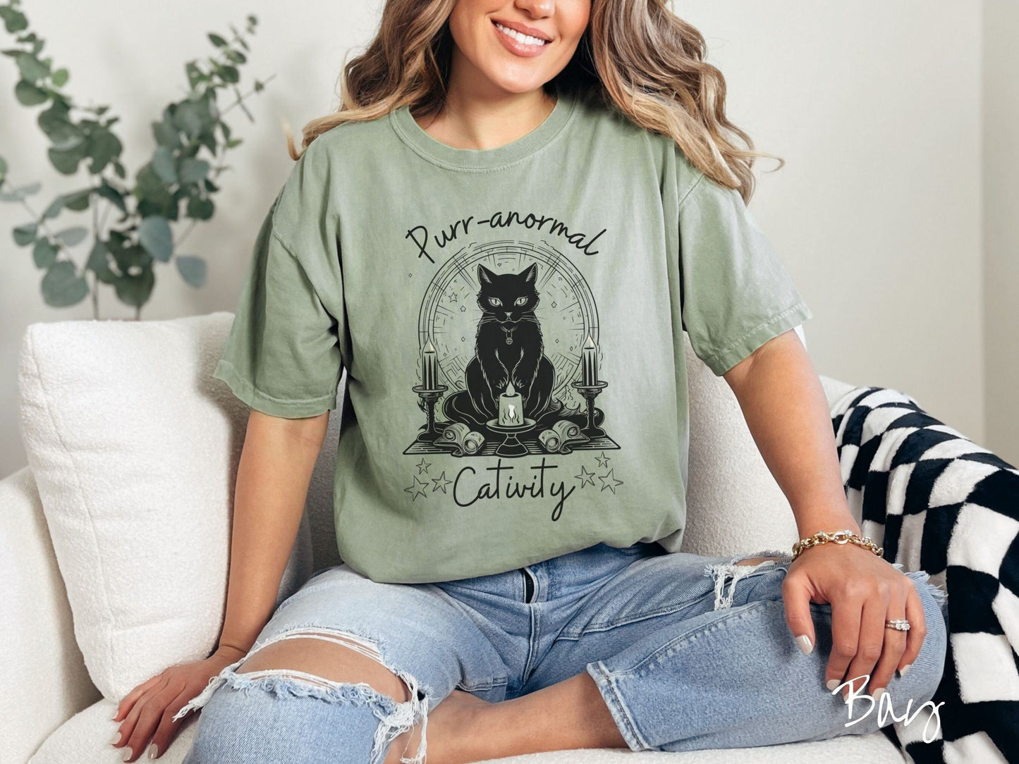 A woman wearing a cute, vintage bay colored Comfort Colors T-shirt with text Purr-anormal Cativity in black font. There is a black cat sitting on an altar with candles and scrolls, staring straight forward with piercing eyes and wearing a necklace.