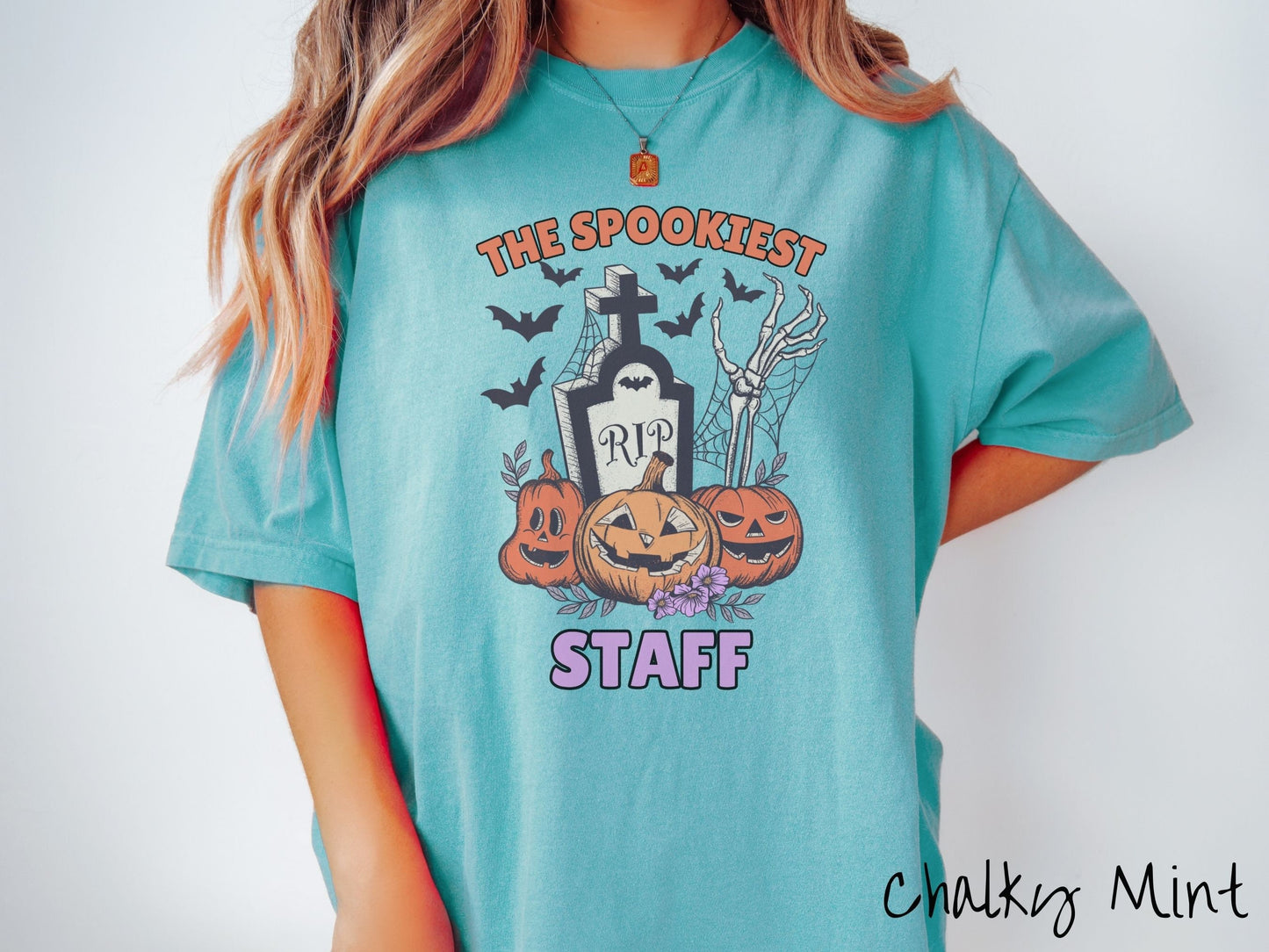 A woman wearing a cute, vintage chalky mint colored Comfort Colors T-shirt with text The Spookiest Staff. There are three smiling carved pumpkins sitting in front of a tombstone with a skeleton arm coming out of the ground and black bats in the air.