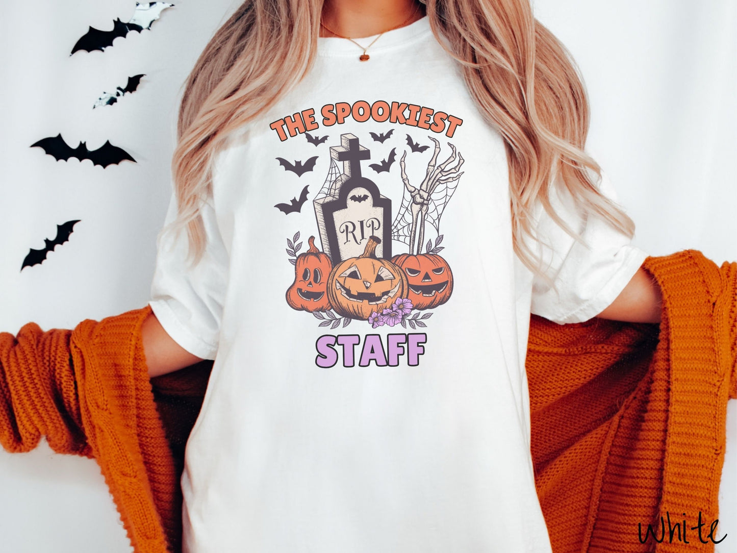 A woman wearing a cute, vintage white colored Comfort Colors T-shirt with the text The Spookiest Staff. There are three smiling carved pumpkins sitting in front of a tombstone with a skeleton arm coming out of the ground and black bats in the air.