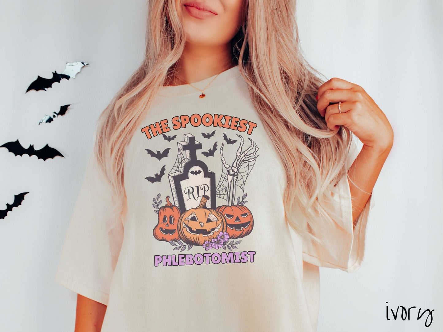 A woman wearing a cute, vintage ivory colored Comfort Colors T-shirt with text The Spookiest Phlebotomist. There are three carved pumpkins sitting in front of a tombstone with a skeleton arm coming out of the ground and black bats in the air.