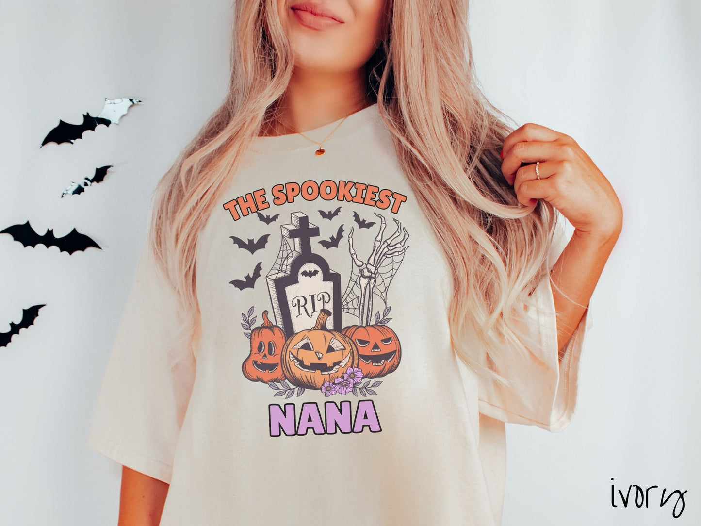 A woman wearing a cute, vintage ivory colored Comfort Colors T-shirt with text The Spookiest Nana. There are three pumpkins sitting in front of a tombstone with a skeleton arm coming out of the ground and bats in the air.