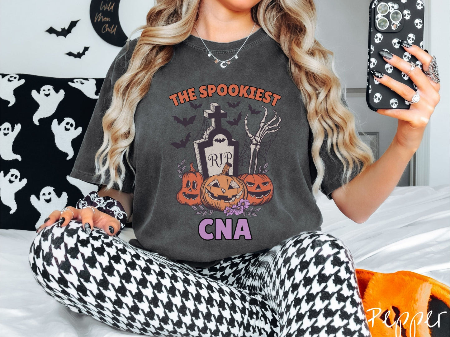 A woman wearing a cute, vintage pepper colored Comfort Colors T-shirt with text The Spookiest CNA. There are three carved pumpkins sitting in front of a tombstone with a skeleton arm coming out of the ground and bats in the air.