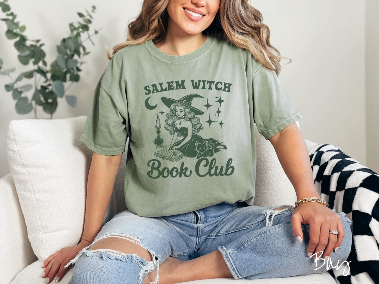 A woman wearing a cute, vintage bay colored Comfort Colors T-shirt with the text Salem Witch Book Club. In between the text is a young female witch reading a book on her knees surrounded by a candle, black cat, and stars and the moon.
