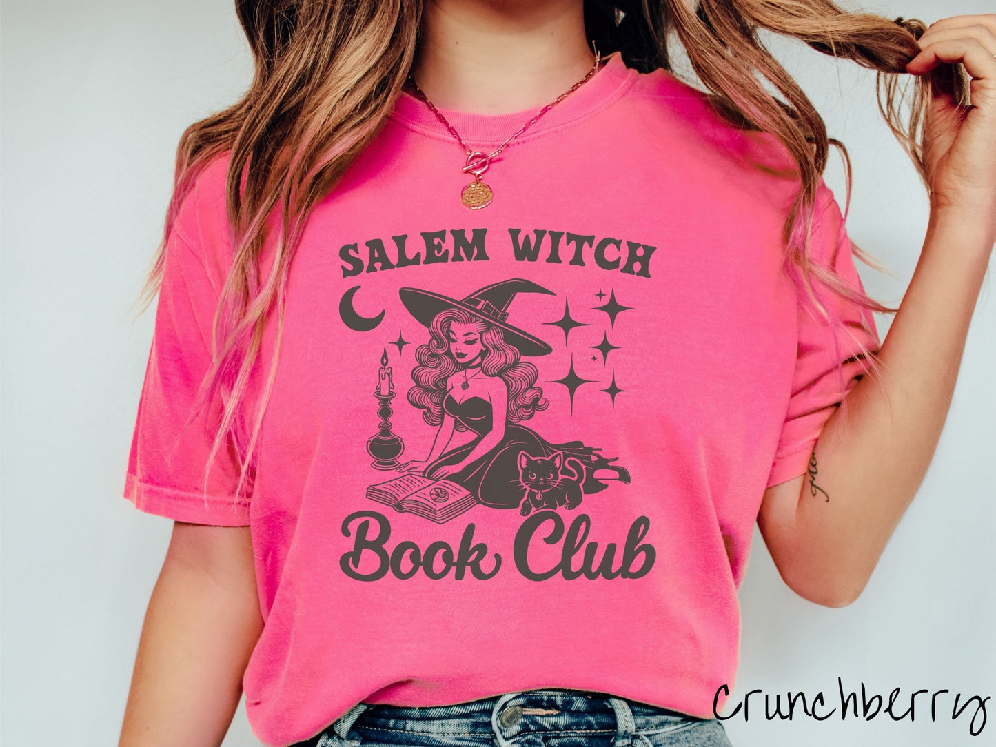 A woman wearing a cute, vintage crunchberry colored Comfort Colors T-shirt with the text Salem Witch Book Club. In between the text is a young female witch reading a book on her knees surrounded by a candle, black cat, and stars and the moon.
