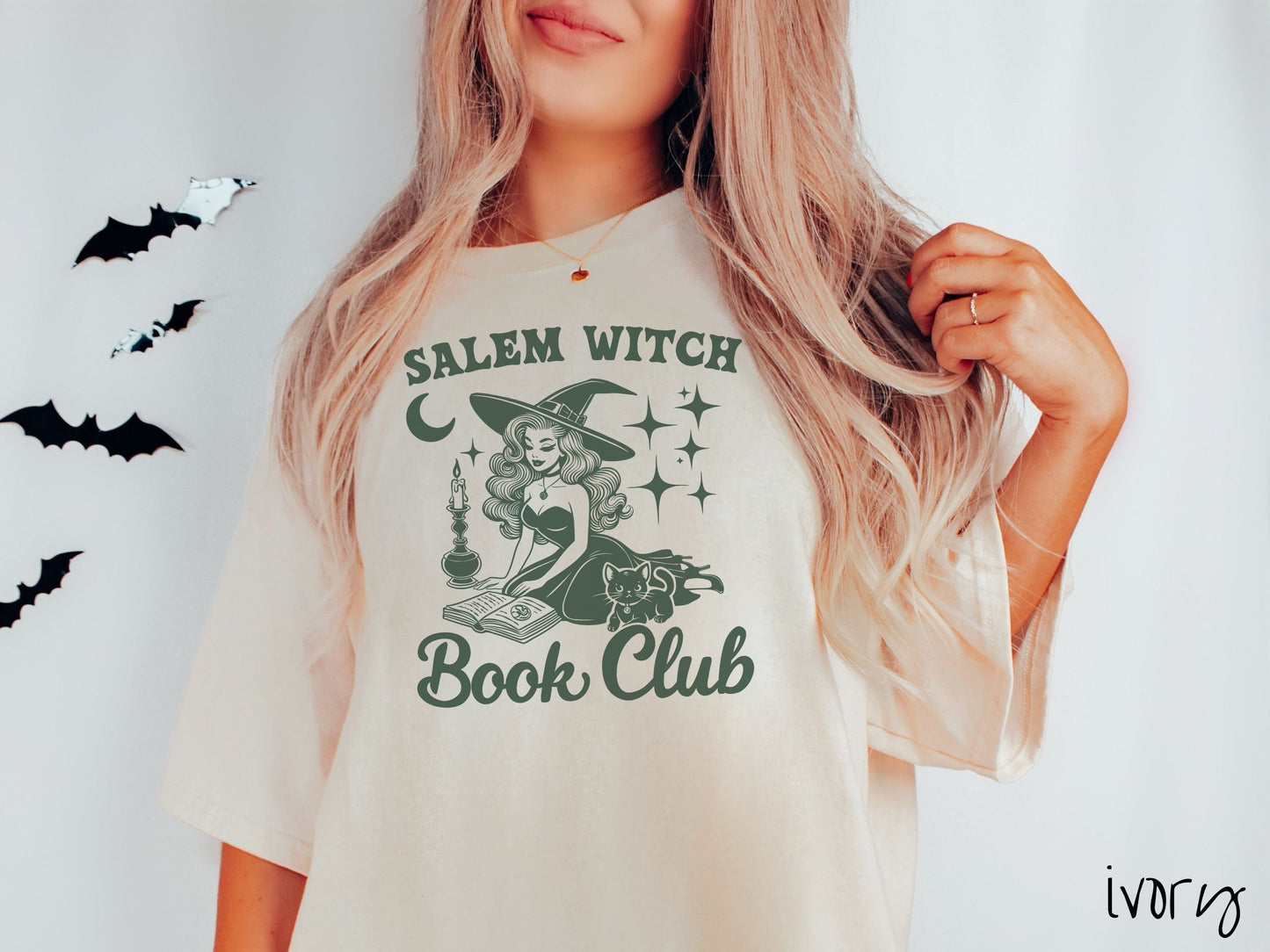 A woman wearing a cute, vintage ivory colored Comfort Colors T-shirt with the text Salem Witch Book Club. In between the text is a young female witch reading a book on her knees surrounded by a candle, black cat, and stars and the moon.