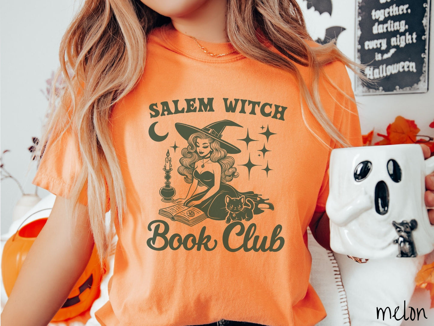 A woman wearing a cute, vintage melon colored Comfort Colors T-shirt with the text Salem Witch Book Club. In between the text is a young female witch reading a book on her knees surrounded by a candle, black cat, and stars and the moon.