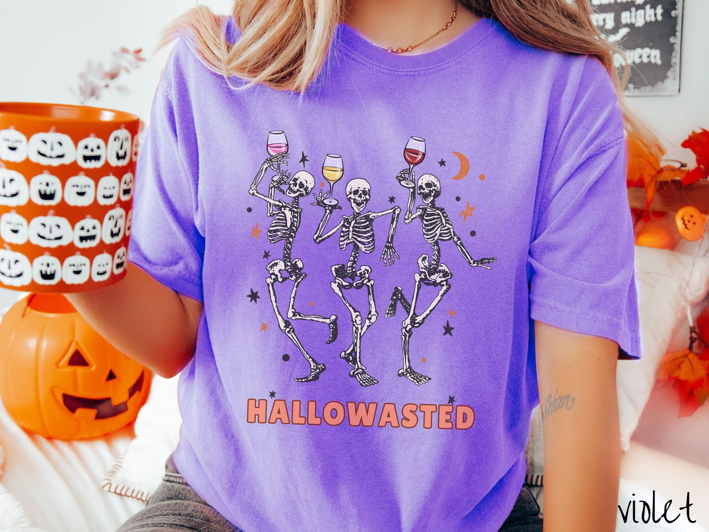 A woman wearing a cute, vintage violet colored Comfort Colors T-shirt with the text Hallowasted in orange font. Above are three female skeletons dancing while holding wine glasses with different colored wines and surrounded by stars.