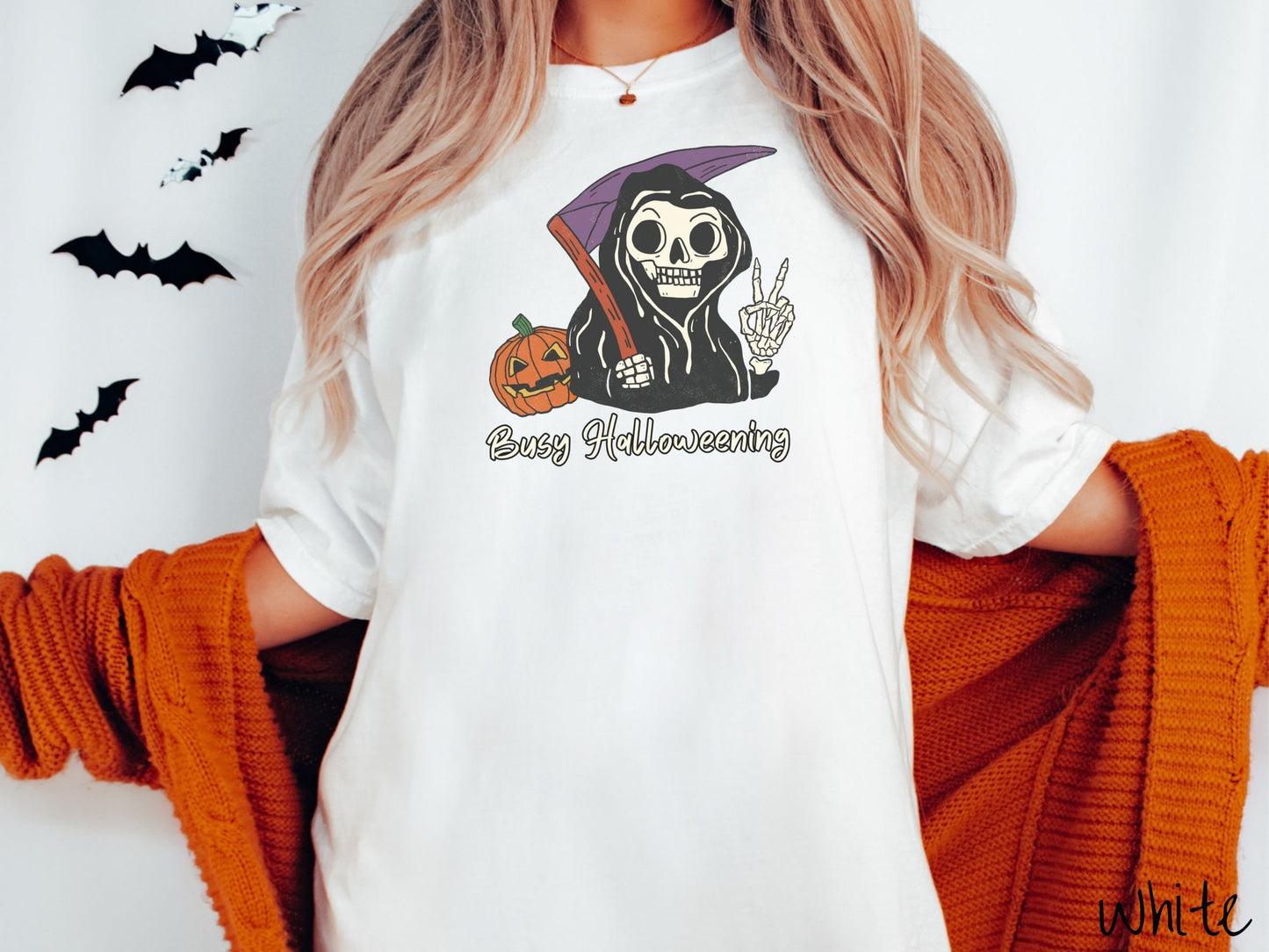 A woman wearing a cute, vintage white colored Comfort Colors T-shirt with text Busy Halloweening in white font. Above is a skeleton wearing a black hood holding up a peace sign in one hand and a scythe in the other with a carved pumpkin behind it.