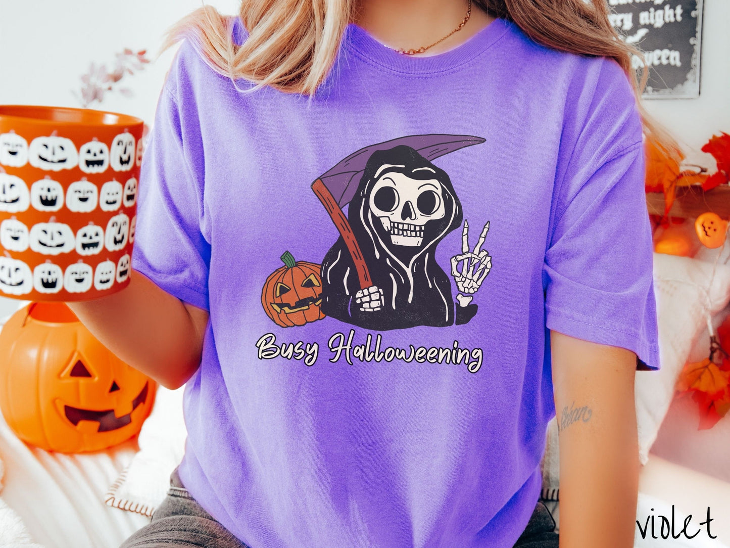 A woman wearing a cute, vintage violet colored Comfort Colors T-shirt with text Busy Halloweening in white font. Above is a skeleton wearing a black hood holding up a peace sign in one hand and a scythe in the other with a carved pumpkin behind it.