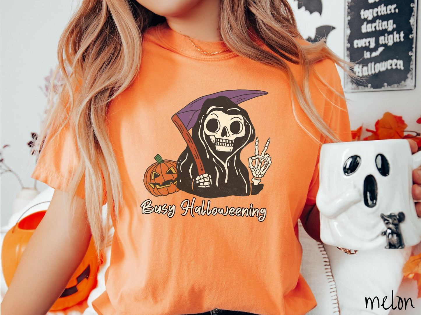 A woman wearing a cute, vintage melon colored Comfort Colors T-shirt with text Busy Halloweening in white font. Above is a skeleton wearing a black hood holding up a peace sign in one hand and a scythe in the other with a carved pumpkin behind it.