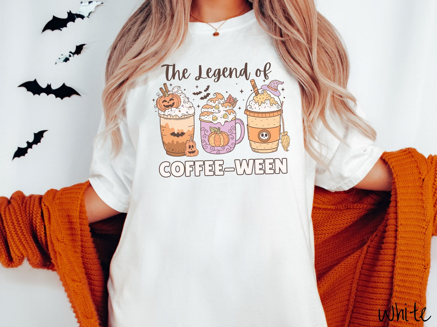 A woman wearing a cute, vintage white colored Comfort Colors T-shirt with the text The legend of Coffee-ween. There are three iced coffees adorned with carved pumpkins, cinnamon sticks, whipped cream, bats, candy corn, whipped cream, and sprinkles.