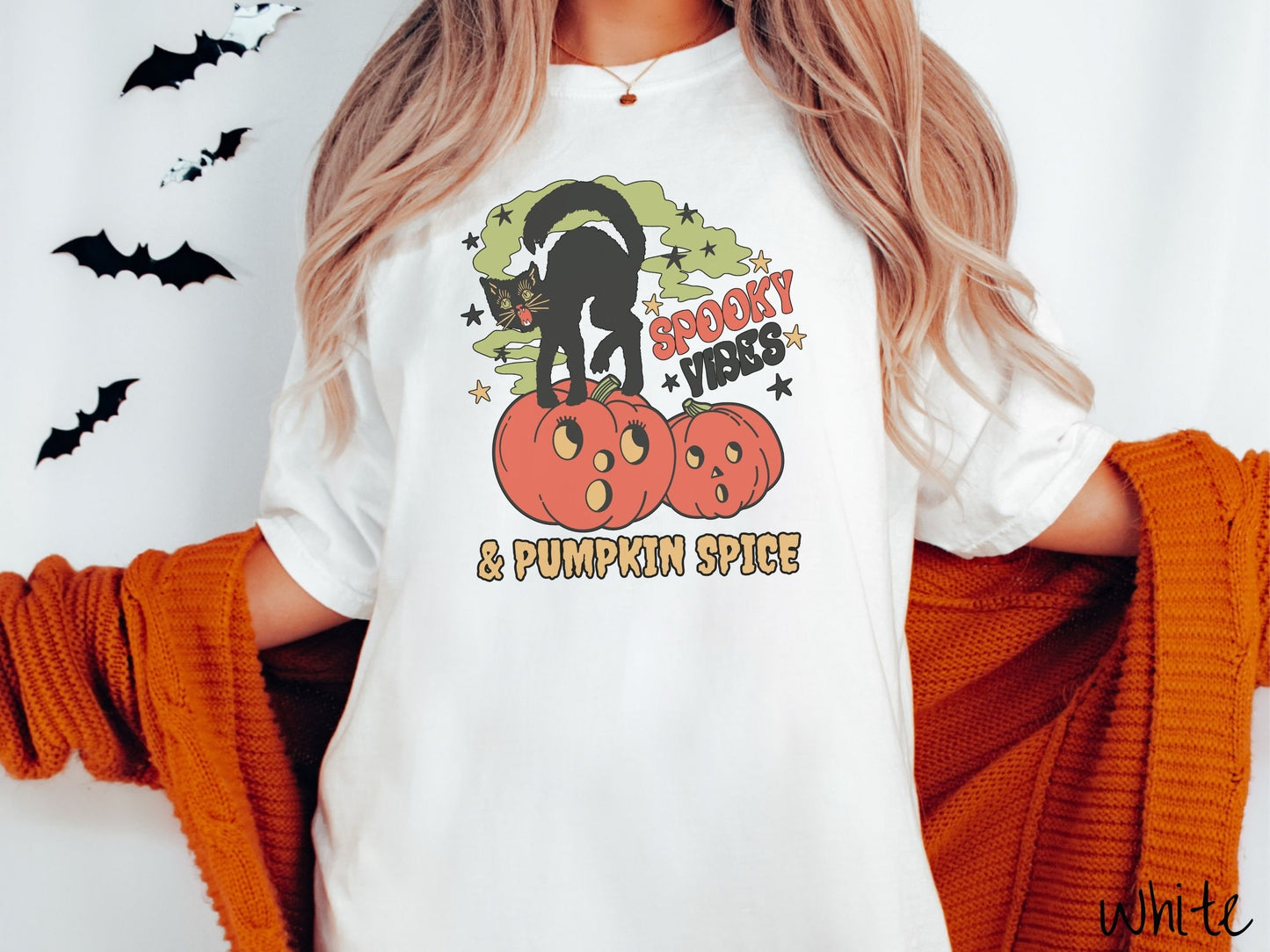 A woman wearing a cute, vintage white colored Comfort Colors T-shirt with the text Spooky Vibes & Pumpkin Spice in orange and black font. There is a black cat standing on an orange carved pumpkin and surrounded by green mist and twinkling stars.