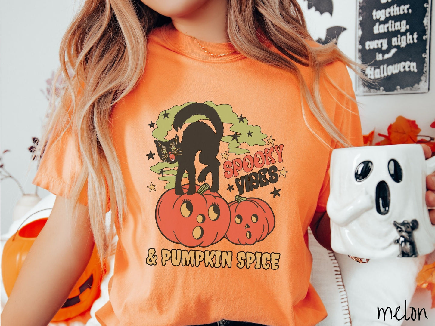 A woman wearing a cute, vintage melon colored Comfort Colors T-shirt with the text Spooky Vibes & Pumpkin Spice in orange and black font. There is a black cat standing on an orange carved pumpkin and surrounded by green mist and twinkling stars.