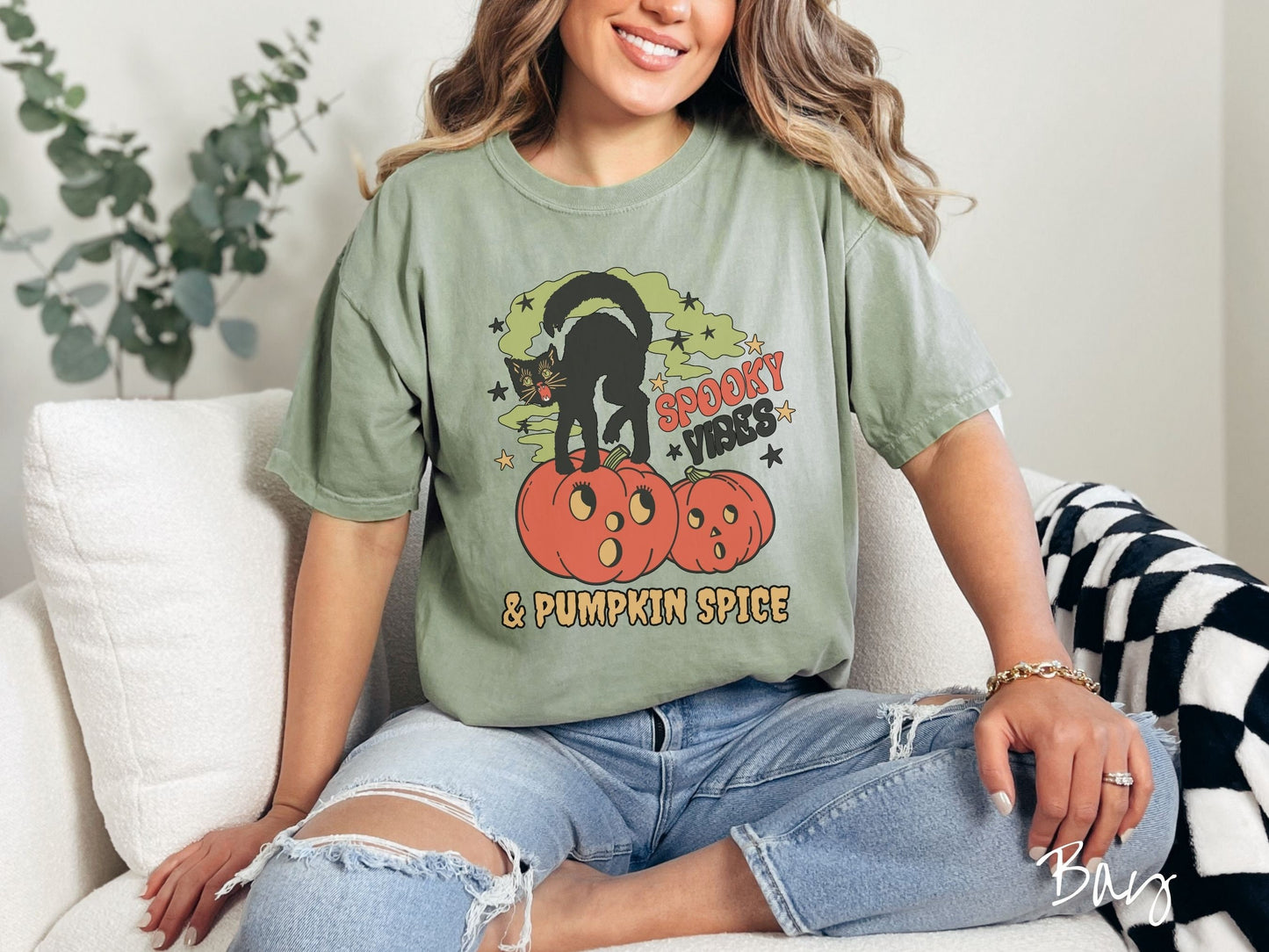 A woman wearing a cute, vintage bay colored Comfort Colors T-shirt with the text Spooky Vibes & Pumpkin Spice in orange and black font. There is a black cat standing on an orange carved pumpkin and surrounded by green mist and twinkling stars.