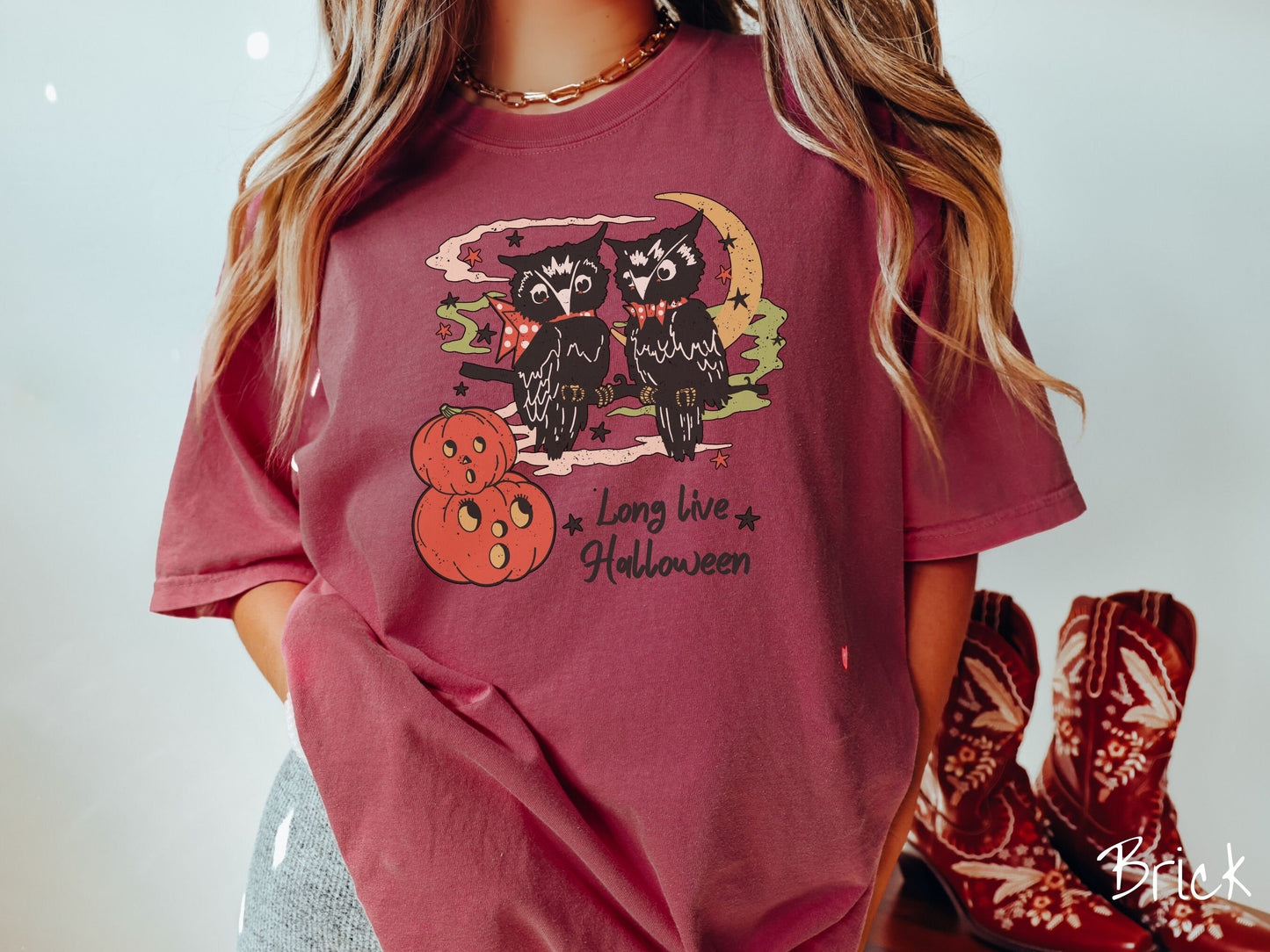 A woman wearing a cute, vintage brick colored Comfort Colors T-shirt with the text Long Live Halloween in black font. Above are two black owls sitting on a tree branch by two scared pumpkins and surrounded by stars and the moon.