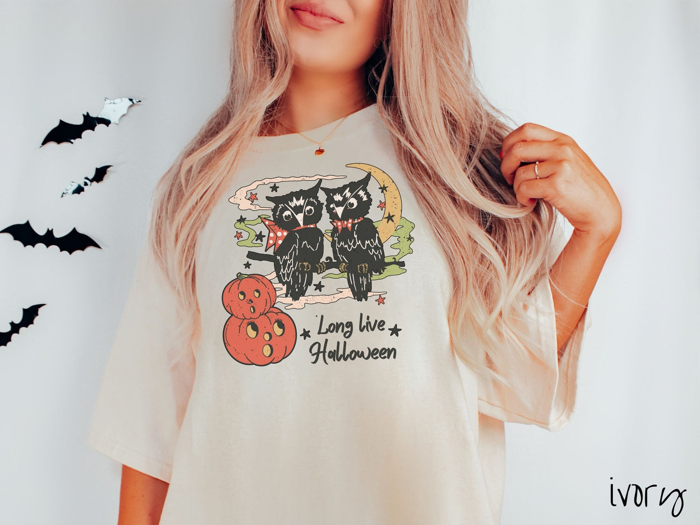 A woman wearing a cute, vintage ivory colored Comfort Colors T-shirt with the text Long Live Halloween in black font. Above are two black owls sitting on a tree branch by two scared pumpkins and surrounded by stars and the moon.
