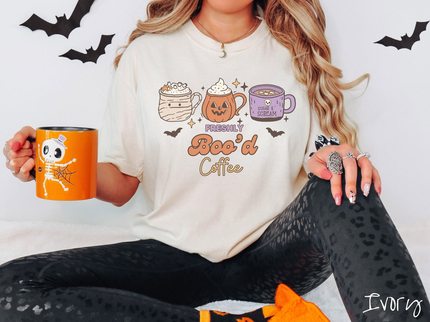 A woman wearing a cute, vintage ivory colored Comfort Colors T-shirt with the text Freshly Boo’d Coffee. Above are coffee mugs where one looks like a mummy with whipped cream, one a carved pumpkin mug, and the last is purple with a skull.