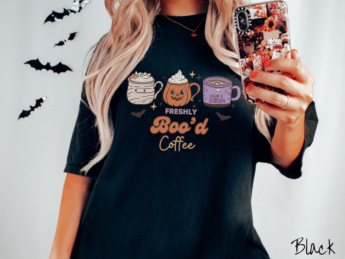 A woman wearing a cute, vintage black colored Comfort Colors T-shirt with the text Freshly Boo’d Coffee. Above are coffee mugs where one looks like a mummy with whipped cream, one a carved pumpkin mug, and the last is purple with a skull.