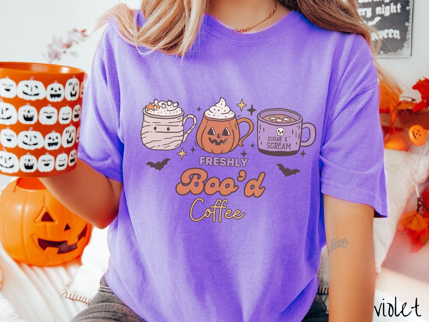 A woman wearing a cute, vintage violet colored Comfort Colors T-shirt with the text Freshly Boo’d Coffee. Above are coffee mugs where one looks like a mummy with whipped cream, one a carved pumpkin mug, and the last is purple with a skull.